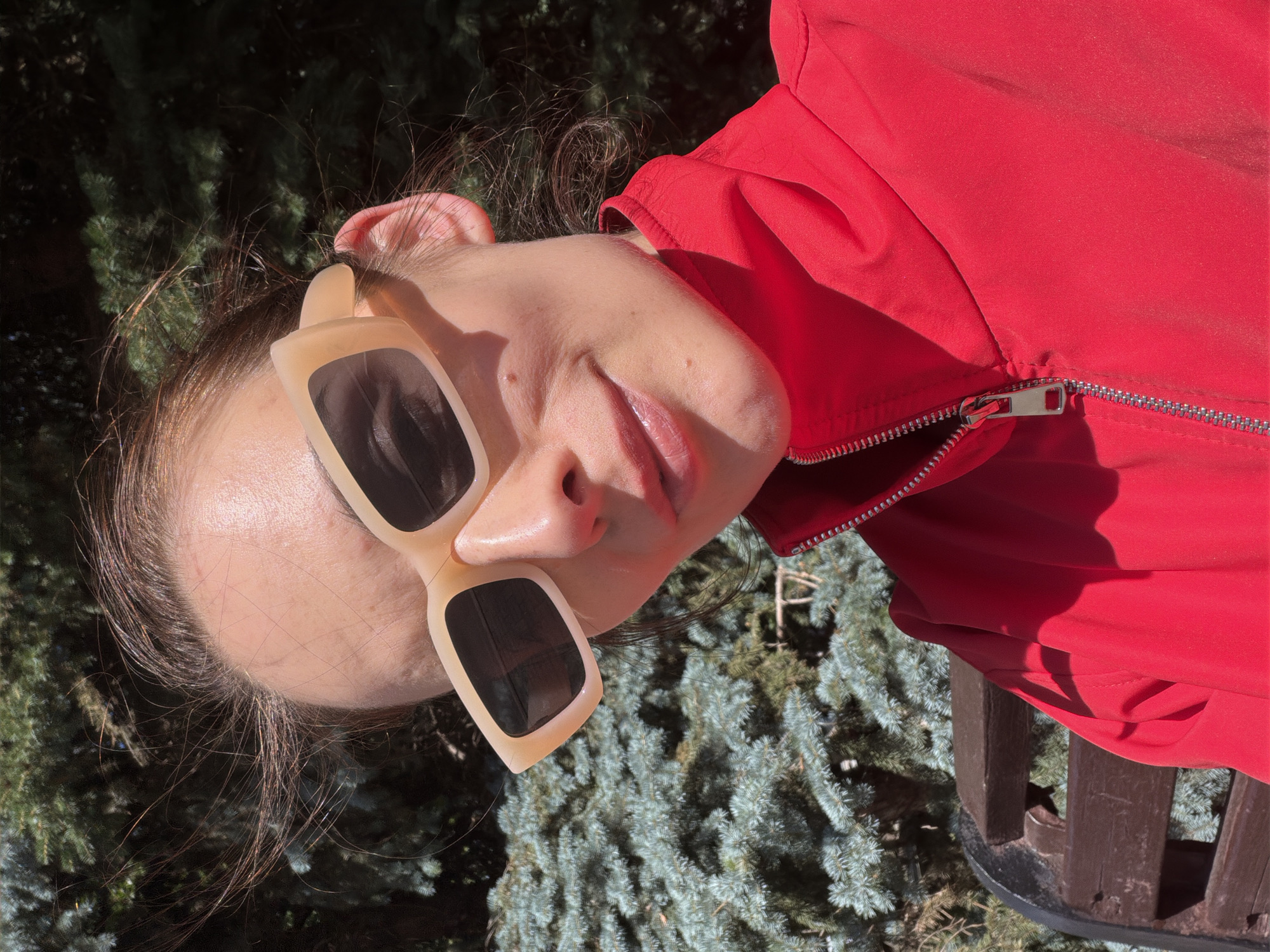 The image shows a close-up, waist-up shot of a young woman wearing oversized, rectangular sunglasses with light-colored frames and a bright red jacket with a zipper. She is outdoors, with pine trees and a wooden bench visible in the background. The lighting suggests it is daytime, with sunlight casting shadows. Her hair is blowing in the wind. The specific location in Ufa, Russia, cannot be determined from the provided image.
