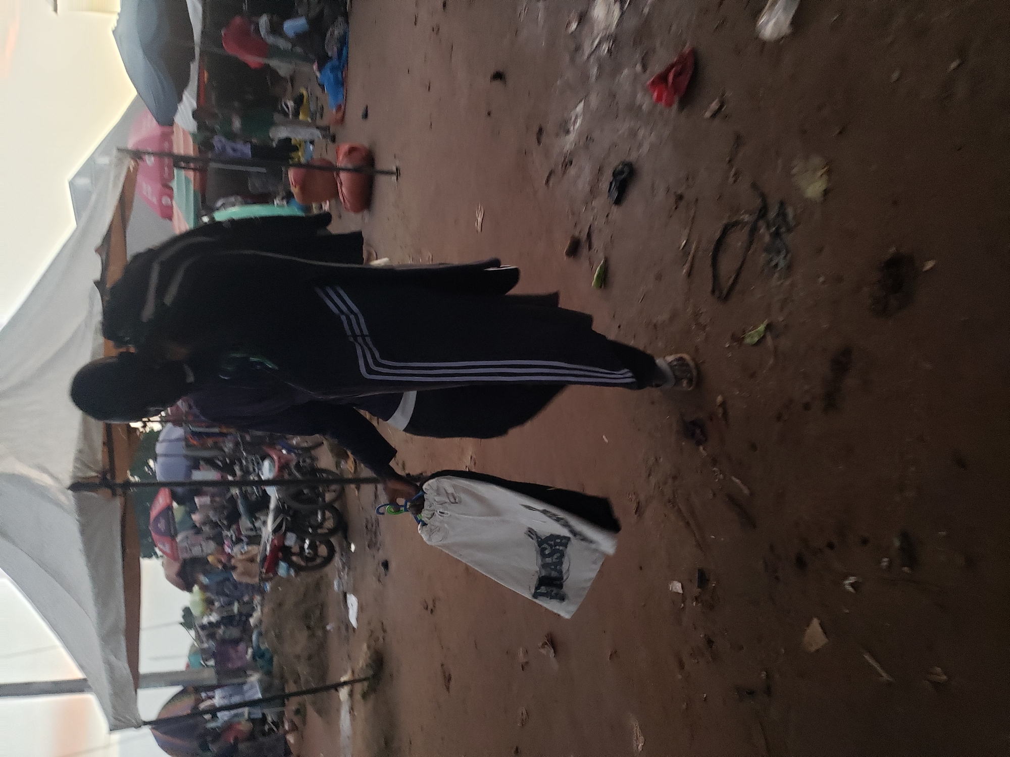 The image depicts an outdoor market scene in Kakuri Gwari, Nigeria. A person, seen from the mid-thigh down, is walking and carrying a white garment on a hanger, which has the word "FLASH" printed on it in black lettering. The person is wearing dark pants with white stripes down the sides and sandals. The ground is a mixture of dry earth, scattered debris, and some muddy patches. In the background, various market stalls with white and colored awnings are visible, along with several motorbikes, people milling about, and some large orange-colored sacks. The overall atmosphere suggests a busy, possibly dusty, marketplace.