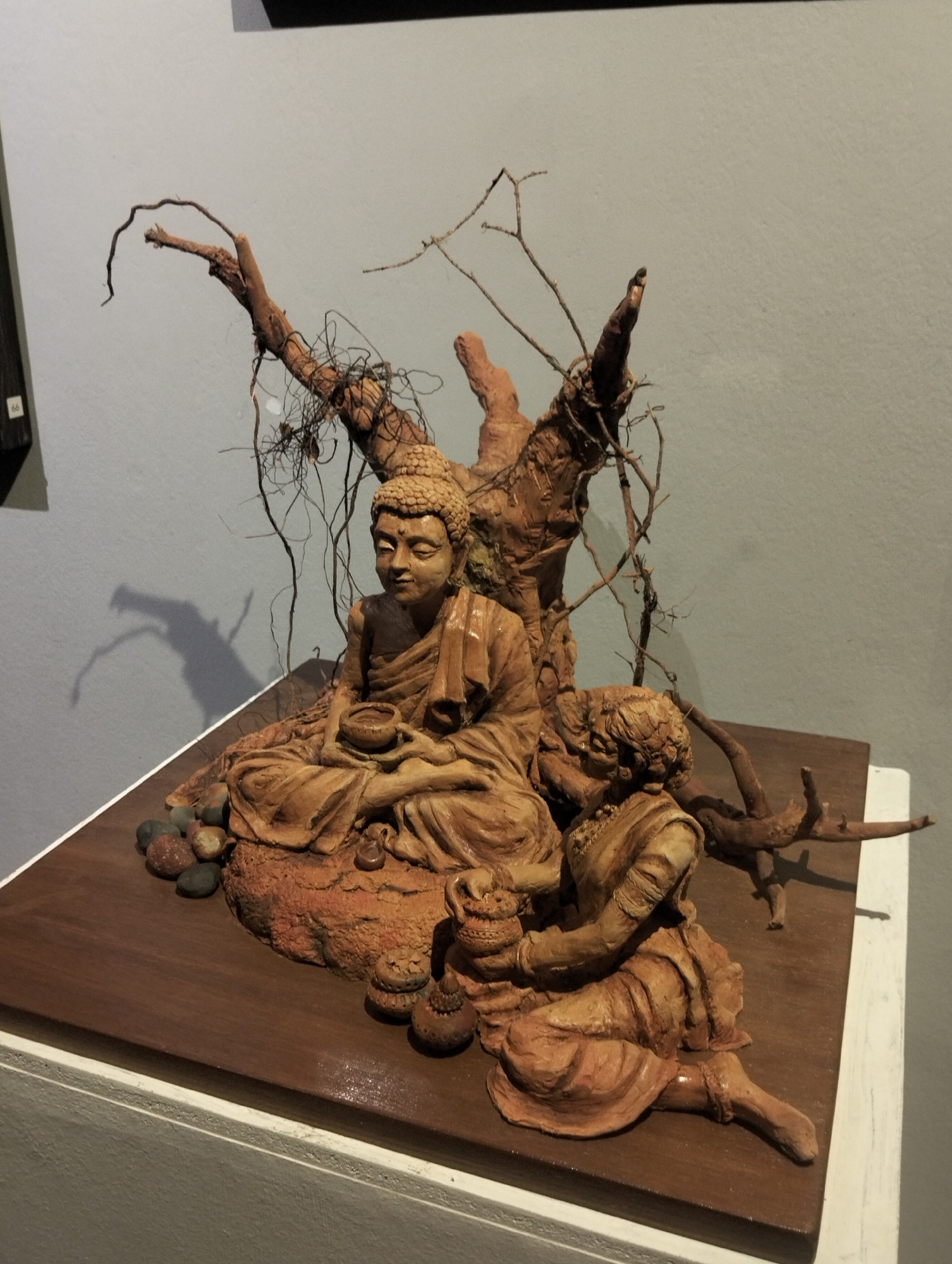 The image depicts a sculpture of Buddha seated in meditation under a tree, accompanied by an attendant. The sculpture is made of a clay-like material and sits on a polished wooden base. The tree behind Buddha is made of branches and twigs. The sculpture is displayed in a gallery or museum setting, likely located in the city of Dhaka, Bangladesh.