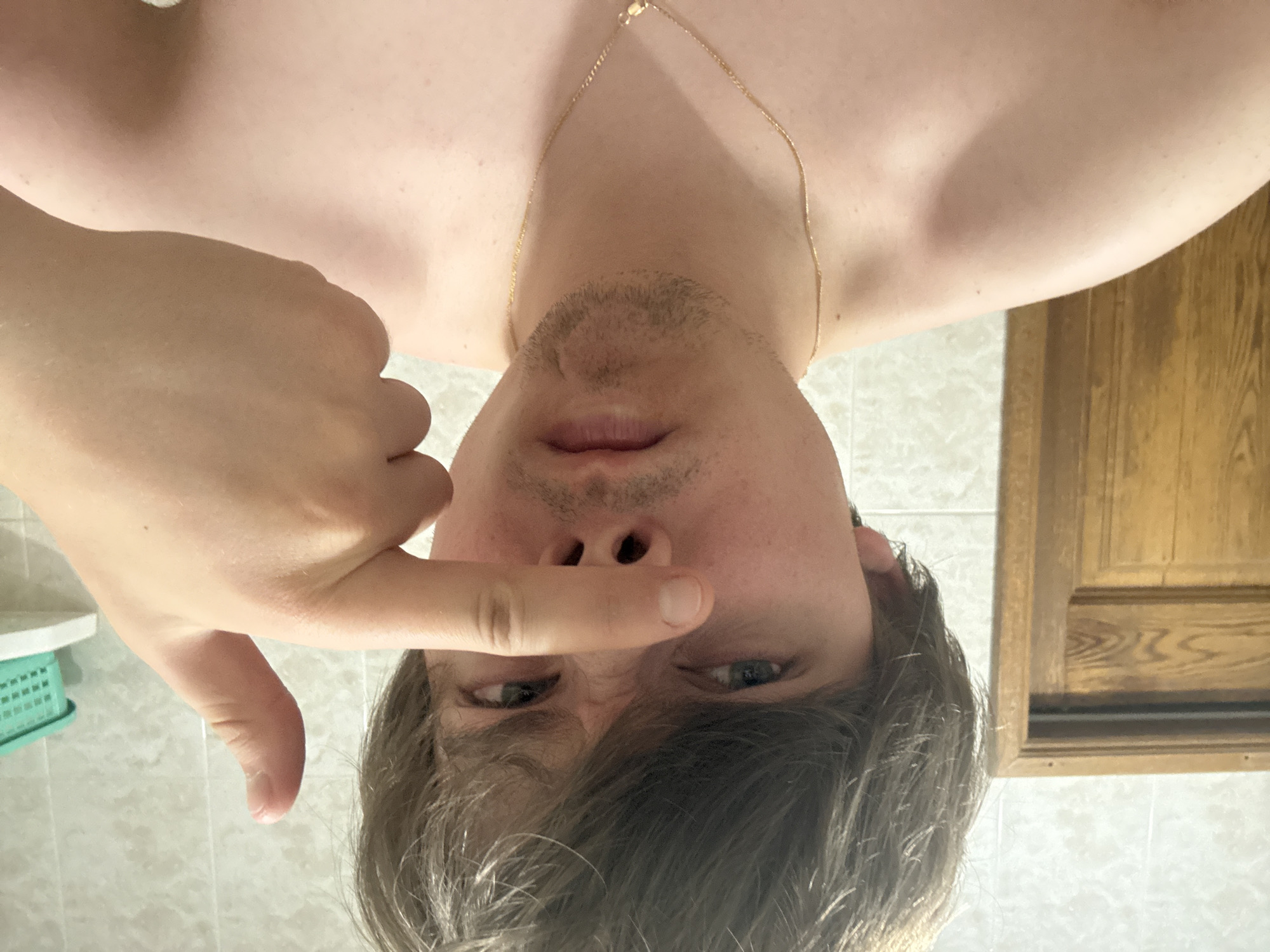 The image shows a close-up of a young person's face and upper chest, taken from a frontal perspective. The person has light skin, light brown hair that is slightly disheveled, and is looking directly at the camera with a playful or mischievous expression. Their right index finger is extended and positioned in front of their nose, with the thumb of the same hand giving a thumbs-up gesture. The person is shirtless, wearing a thin gold chain around their neck. The background is a tiled wall, possibly in a bathroom, with a wooden door on the left and a shelf with a green plastic basket on the right. There is no discernible information in the image that would allow for the determination of the location in the city of Dnipro, Ukraine.