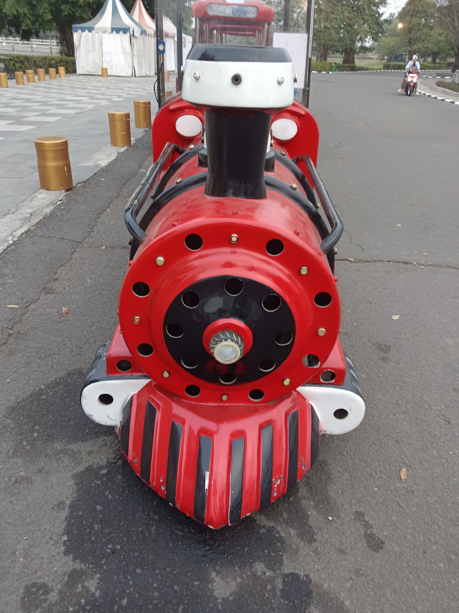 In the city of Jayamukti, Indonesia, a toy train sits on an asphalt surface. The train is predominantly red with black and white accents. It features a large, circular front with multiple circular cutouts and a central mechanism. Black and red stripes adorn the lower front of the train. To the left, golden cylindrical posts are lined up on a tiled walkway, with tents in the background. In the distance, on the asphalt road, a person is riding a motorcycle. The environment suggests an outdoor recreational area, possibly a park or a public space.