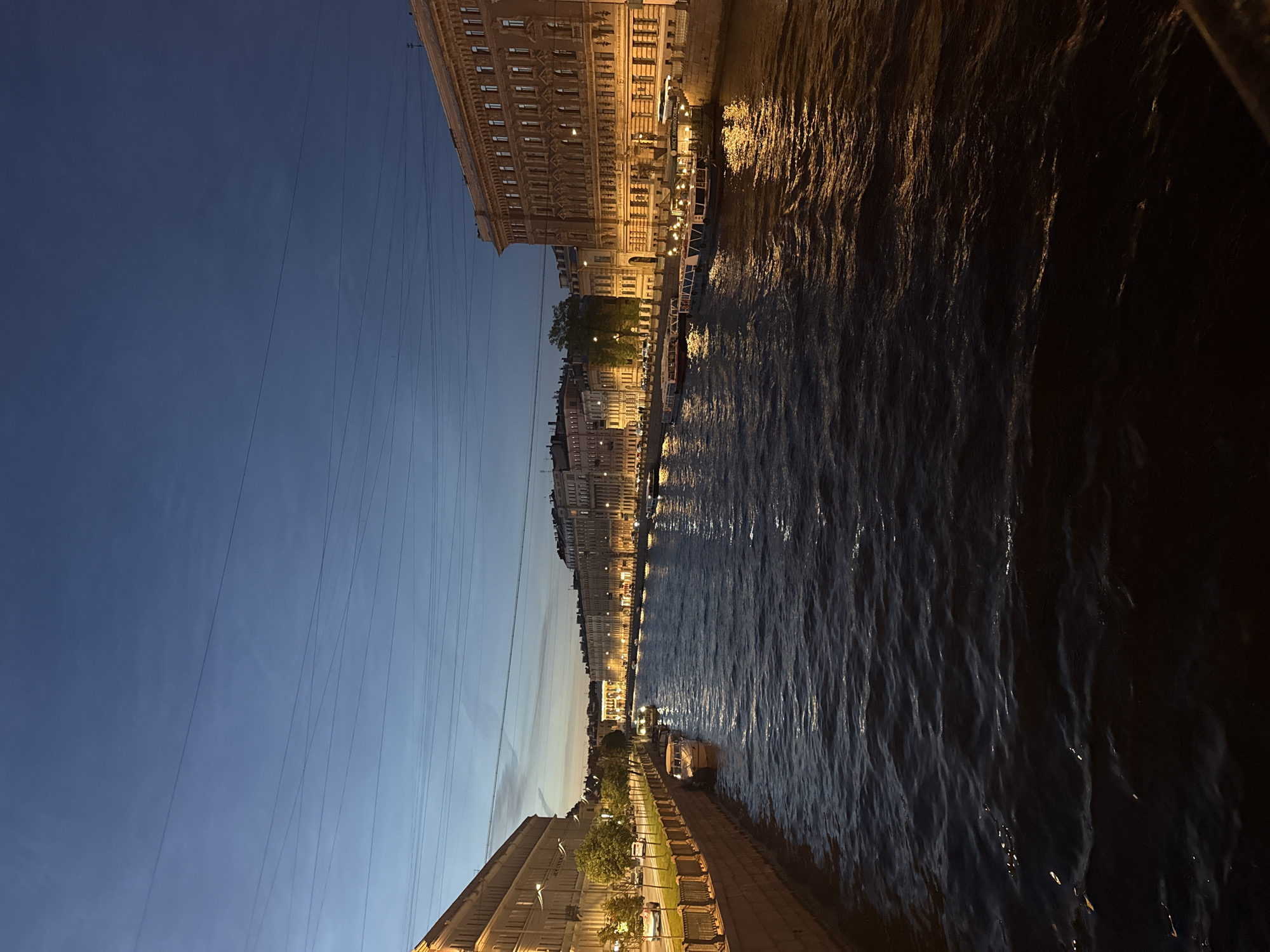 This image depicts a wide, dark canal or river in Saint Petersburg, Russia, viewed from a bridge or elevated vantage point. The scene is set during twilight, with the sky transitioning from a deep blue to a lighter hue near the horizon, and a few faint wires stretch across the upper portion of the frame.
Along both banks of the waterway are multi-story buildings with illuminated windows, suggesting a densely populated urban area. These buildings are characterized by classical architecture, with ornate facades and repeating window patterns. Along the left bank, trees and a sidewalk with a decorative fence are visible, with a few cars driving on the street. Several boats are docked along the canal, some appearing to be passenger vessels with lights on, hinting at evening excursions. The water itself is dark and rippled, reflecting the warm glow of the lights from the buildings and boats, creating streaks of yellow and orange on its surface. No people are clearly discernible in the image. The overall atmosphere is serene and urban, characteristic of a European city at dusk.