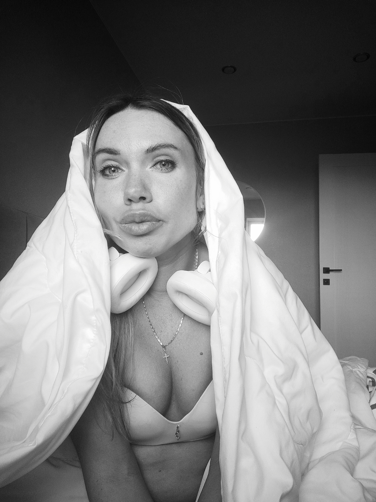 A grayscale medium shot depicts a woman indoors. She is draped in a white blanket over her head, shoulders, and chest. She wears white over-ear headphones around her neck and a necklace with a cross pendant. She is also wearing a white bra. She is looking directly at the camera with a slightly puckered expression. The background includes a wall, a door, and a round mirror. The location is unknown and there is no information to suggest that the photo was taken in Votkinsk, Russia.