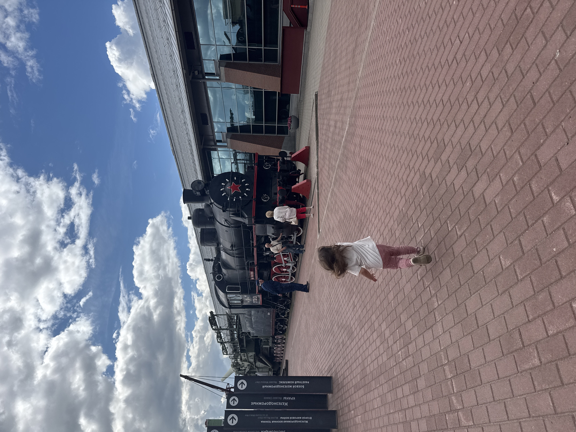 In Saint Petersburg, Russia, at the Central Naval Museum, a young child is captured in motion, running on a red brick pathway. The child, wearing a white t-shirt and pink pants, is facing away from the camera. In the background, a large black steam locomotive stands prominently on display, adorned with a red star emblem. Several adults are visible near the locomotive, observing the exhibit. Behind the train, a modern building with large glass windows reflects the sky and clouds. The environment suggests an outdoor exhibition space with signage posts visible to the left of the frame. The sky is partly cloudy, with a mix of blue sky and white cumulus clouds.