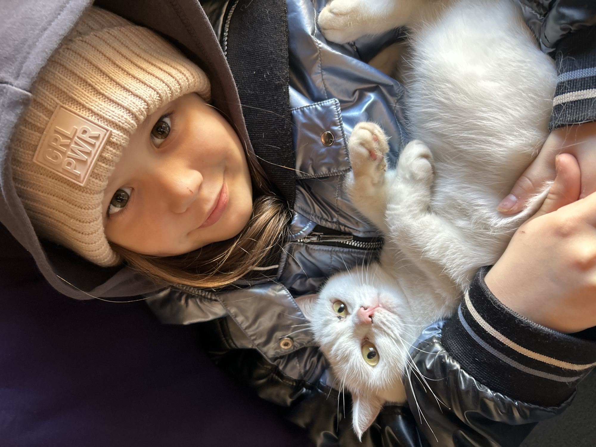 A young girl in a beige knit cap with "GRL PWR" printed on a square patch on the front and a gray hooded jacket is lying on her back and holding a white cat on her chest. The girl is smiling at the camera. The cat is lying on its back with its paws in the air. The scene is illuminated by sunlight. The location is Chaikovsky, Russia.