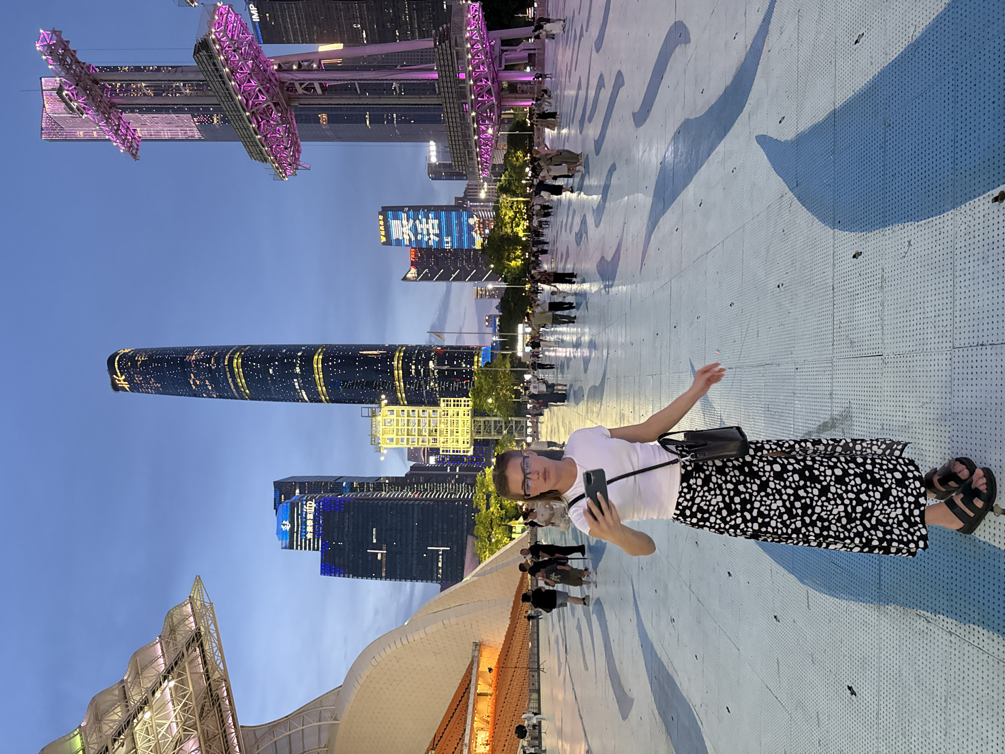The image depicts a woman taking a selfie with a smartphone in an urban plaza at dusk. She is wearing a white t-shirt, a black and white patterned skirt, and black sandals, with a black purse slung over her shoulder. The plaza floor has a blue and white wavy pattern.
In the background, a cluster of modern skyscrapers are illuminated. One tall, slender skyscraper is particularly prominent, featuring a spiral design and numerous lit windows. Other buildings display bright lighting, including one with blue and white signage. Above, a large structure with purple lighting, possibly part of a Ferris wheel or observation tower, is visible against the darkening sky. Several other people are scattered throughout the plaza, appearing to be walking or gathering.
The time of day is evening, as indicated by the twilight sky and the illuminated buildings. The weather appears to be clear. The location is identified as Tianhe District, China, a known hub for modern architecture and urban development. The woman's pose and the selfie suggest she is enjoying her surroundings and documenting her experience. The overall scene conveys a sense of a lively urban evening.