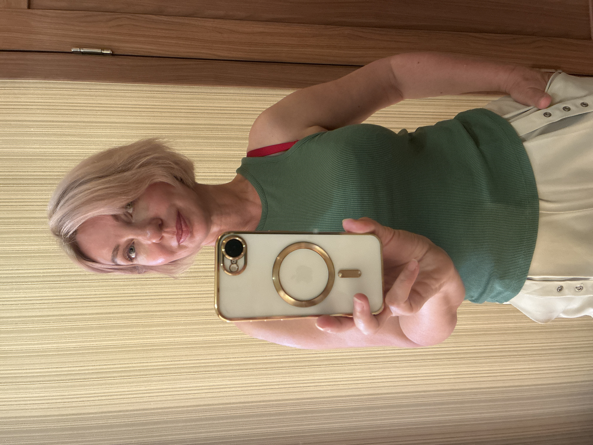 A middle-aged female with short, blonde hair and light-colored eyes is depicted taking a mirror selfie indoors in Sochi, Russia. She is wearing a ribbed, green tank top, with a red bra strap visible on her left shoulder. Her lower attire consists of light-colored trousers or shorts featuring metallic snap closures along the outer seam of the pockets, with her right hand partially inserted into the pocket. She holds a light-colored smartphone with a gold-trimmed case, which includes a gold-colored MagSafe ring and camera accent, in her left hand, positioned centrally in front of her. The background consists of a textured, vertically striped, light beige wallpaper on the left, and a light-colored wooden door with a visible silver hinge on the right.