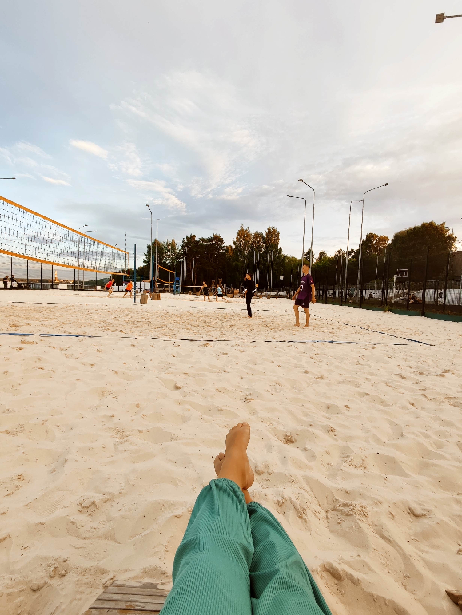 The image depicts a beach volleyball court on a sandy surface under a cloudy sky. Several individuals are engaged in a game of volleyball. On the far left, a net divides the court. In the foreground, a person's legs and feet are visible, suggesting they are seated or lying down on the sand, observing the game. Further into the court, players in athletic attire are actively participating in the game, some running, others in positions to hit or receive the ball.
The lighting suggests it is either early morning or late afternoon, with diffused sunlight filtering through the clouds. The overall atmosphere appears casual and recreational. Trees form a backdrop to the court, indicating an outdoor setting, possibly a park or a dedicated sports complex. There are no visible signs or text in the image. The focus is on the beach volleyball activity and the serene, yet active, scene.