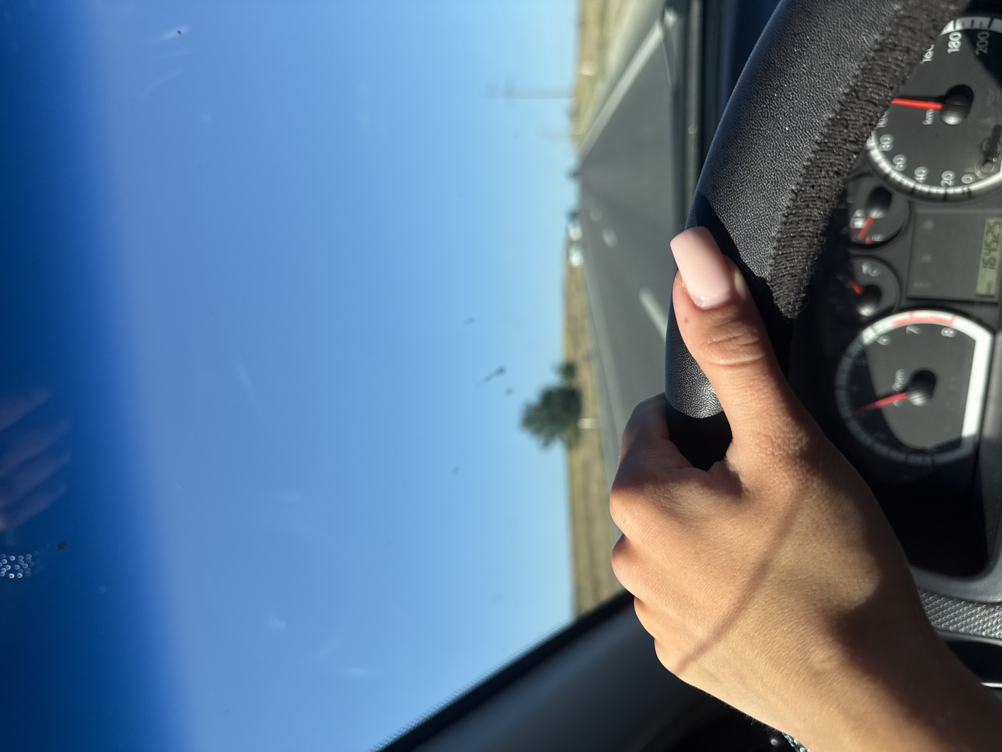 The image shows a close-up, interior view of a person's right hand gripping a black steering wheel of a car. The hand has neatly manicured nails painted a light pink color. The dashboard is partially visible, with a speedometer showing a speed between 80 and 100 km/h. Outside the car, a bright blue sky is visible through the windshield, with a straight, paved road stretching into the distance. The road is bordered by dry, grassy terrain. Several other vehicles are visible as blurred shapes on the road ahead. The scene appears to be a sunny day. There is no information to suggest the location is Comrat, Moldova.