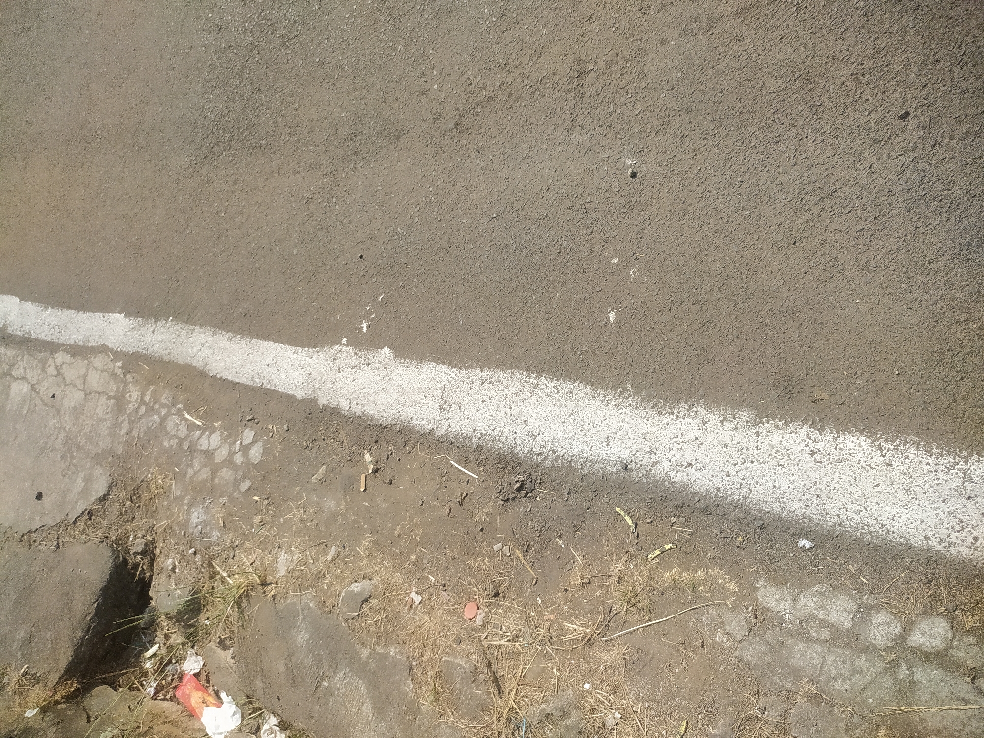 An outdoor, eye-level, medium shot captures a section of an asphalt road adjacent to a dirt embankment. A thick, white line demarcates the edge of the road. The embankment is comprised of soil, small rocks, and dry grass. Litter, including plastic wrappers and debris, is visible scattered on the embankment. The road surface is a granular asphalt. The scene is illuminated by natural sunlight, casting subtle shadows. The environment suggests a rural or roadside setting within the city of Lopait, Indonesia. No people or animals are present.
