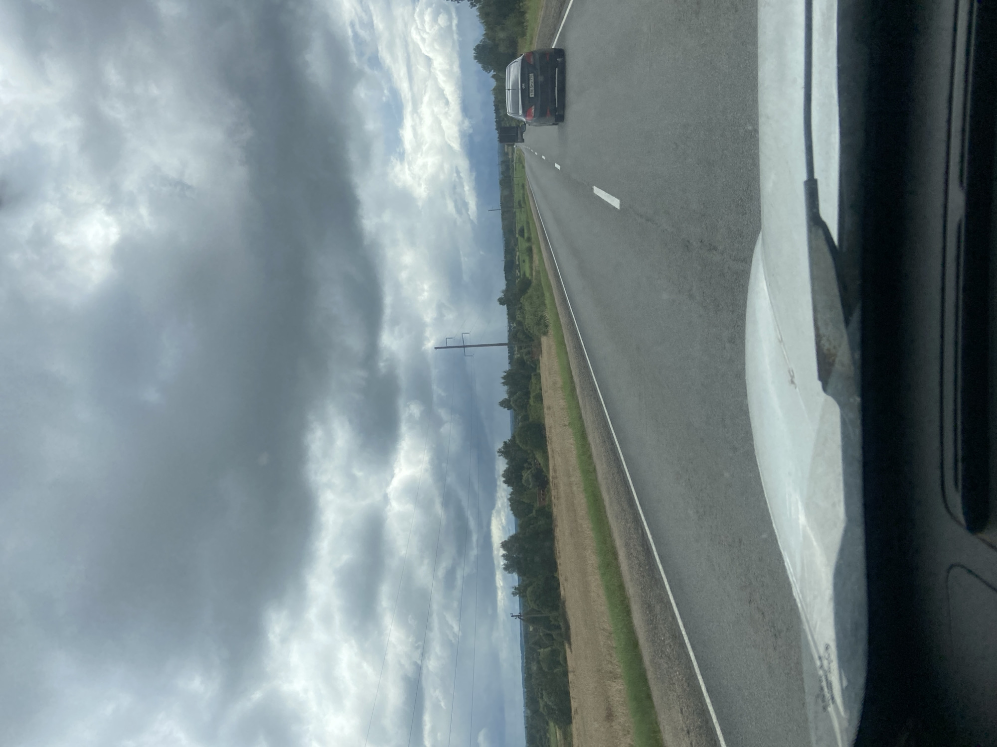 The image depicts a view from inside a vehicle traveling on a paved road. The road has white lane markings and is bordered by a grassy shoulder. On the left side of the road, there is a field of dry grass or crops, followed by a line of trees and utility poles against a cloudy sky. On the right side of the road, a dark-colored sedan is visible in the distance. The sky is filled with heavy, gray clouds, suggesting overcast weather. The foreground shows the dashboard and windshield of the vehicle, with some reflections on the glass. The location is specified as Заячий-Холм, Russia.