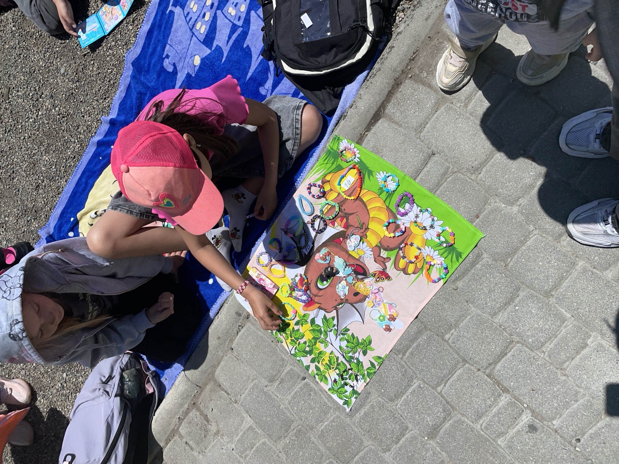 A high-angle shot reveals a group of people, primarily children, gathered on a sunny outdoor surface, likely a sidewalk in Vladivostok, Russia. The setting appears to be urban, with interlocking grey brick pavers forming the ground.
A girl in a pink baseball cap, shorts, and socks with Mickey Mouse imagery kneels beside a blue towel laid on the ground. Next to her is a colorful cartoon-themed cloth or mat covered with various handmade bracelets. She is reaching toward the mat, presumably examining or selecting a bracelet.
Another child, clad in a grey hooded sweatshirt, sits on the towel, appearing to observe the scene. Scattered around are personal belongings, including backpacks and jackets.
Additional individuals stand nearby, partially visible in the frame, their feet suggesting adults present. The lighting is bright, casting shadows and indicating a clear, sunny day. The atmosphere suggests a casual outdoor gathering, possibly a small impromptu market or play session.