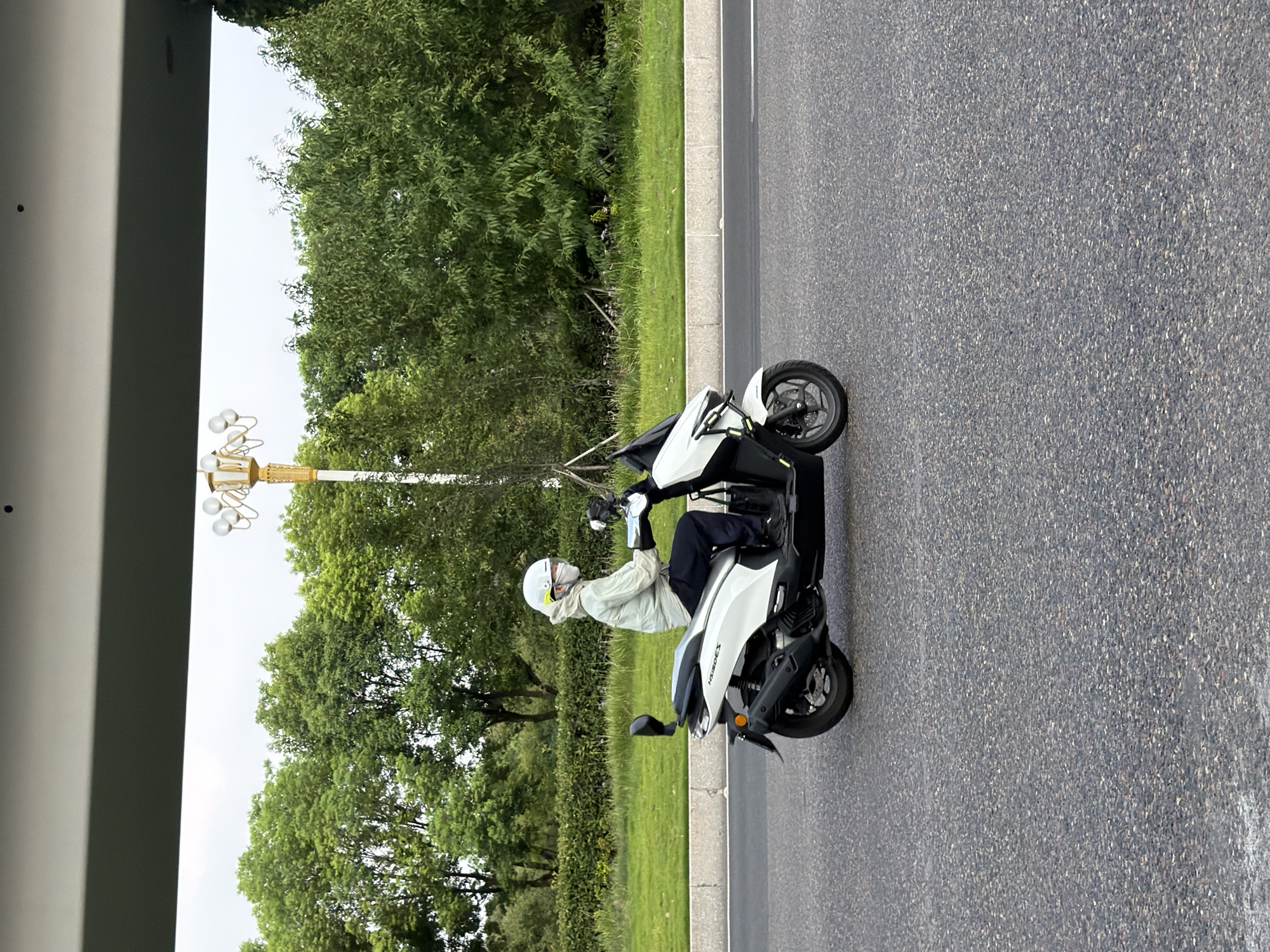 A person wearing a white helmet and a light-colored hooded jacket rides a white scooter on an asphalt road. The scooter has black accents and the model name "NEXGEX" is visible on its side. The person is wearing dark pants and black shoes. In the background, there is a grassy area with a hedge and lush green trees. A decorative lamppost with multiple light fixtures stands behind the scooter. A gray overhang or structure is visible in the top portion of the image. The scene appears to be in a city park or along a tree-lined street.