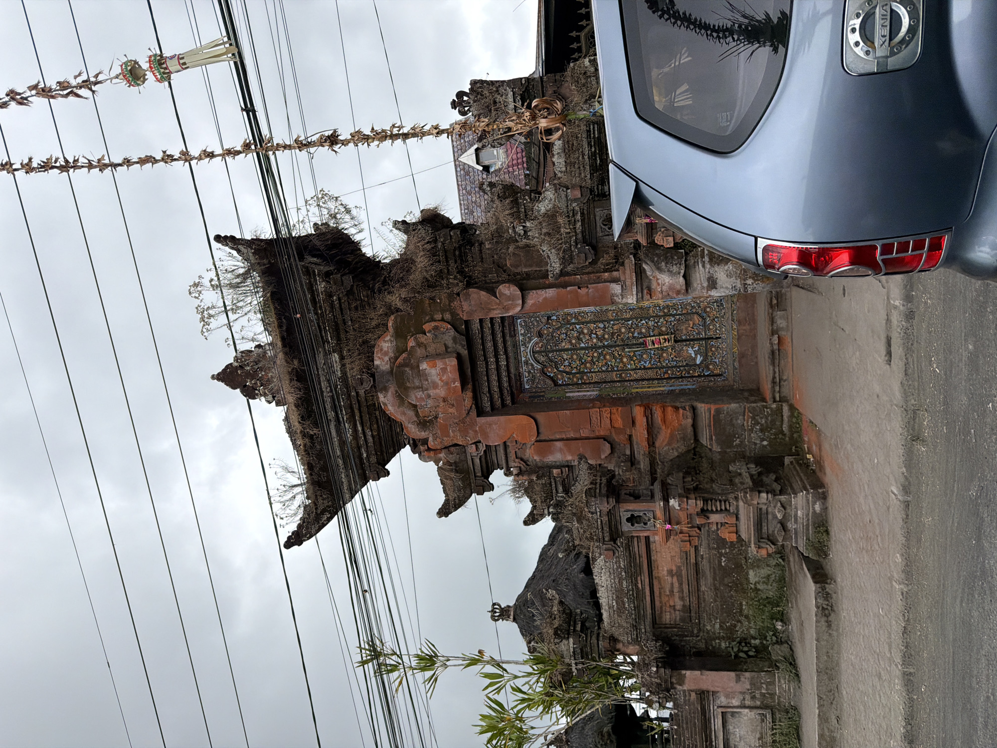 The image depicts a street scene in Kintamani, Indonesia. In the foreground, the rear passenger side of a light blue SUV is visible, showing its taillight and "XENIA" badging. The vehicle is parked on an asphalt road. Behind the car is a traditional Balinese temple gate, constructed from reddish-brown brick and stone. The gate is elaborately carved and adorned with moss and overgrowth, suggesting age and a humid climate. Above the gate, numerous electrical wires crisscross the sky, which is overcast and gray. Two decorative strings of dried leaves and paper ornaments hang from the wires, one of which is more ornate with red and white details. A small, leafy bamboo plant grows to the left of the gate. The overall atmosphere is somewhat weathered and tropical.