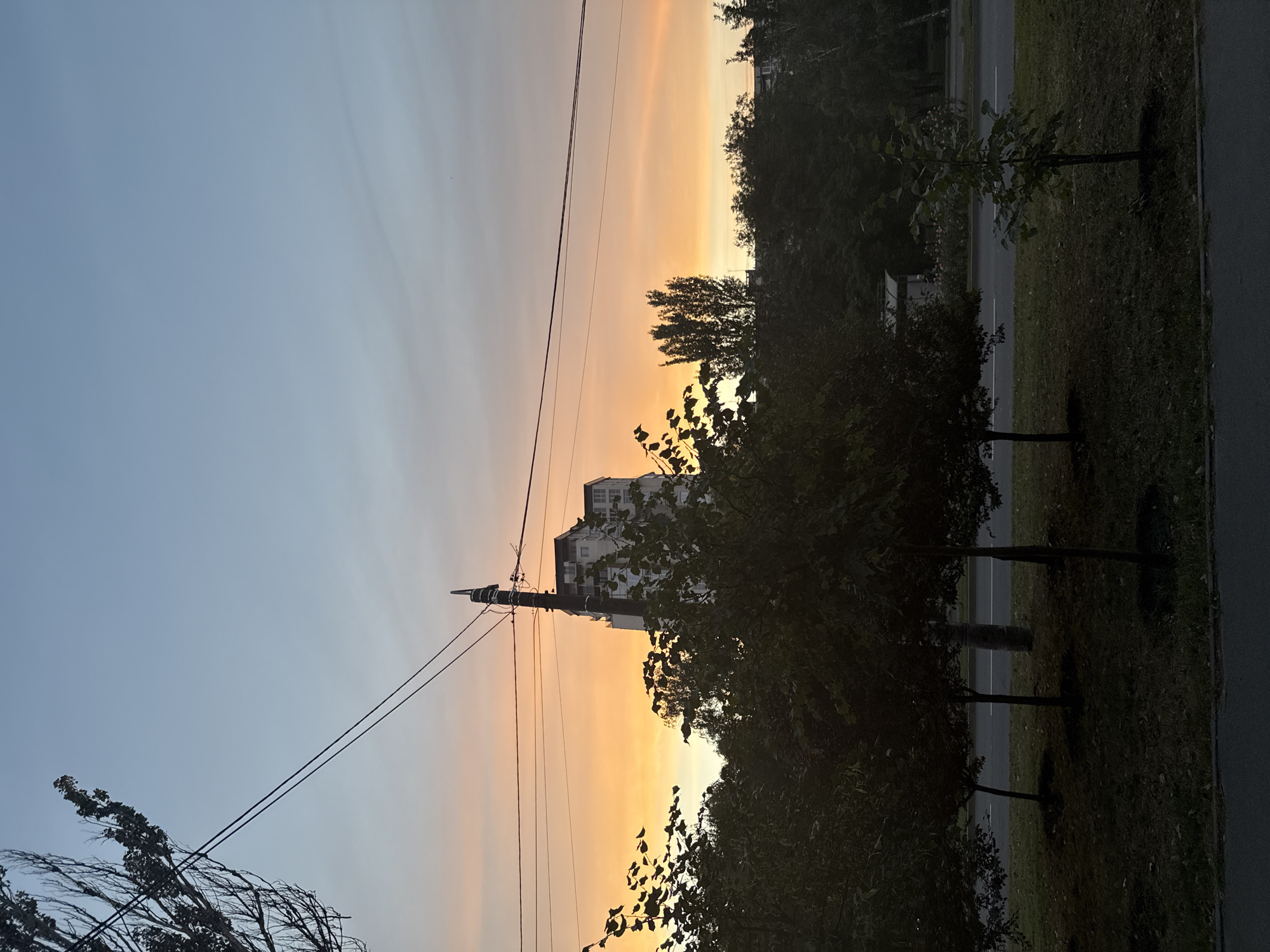 The image depicts a street scene in Zaporizhzhia, Ukraine during a sunset. The sky is a gradient of light blue and orange, with wispy clouds. In the background, a tall, multi-story building with a dark roof stands prominently. Electrical wires and a utility pole run across the frame, obscuring parts of the building and the sky. In the foreground, several trees with dark green foliage are silhouetted against the brighter sky. The lower part of the image shows a paved road with a grassy median strip where young trees are planted. The overall impression is one of an urban landscape transitioning into evening. There are no people visible in the image.