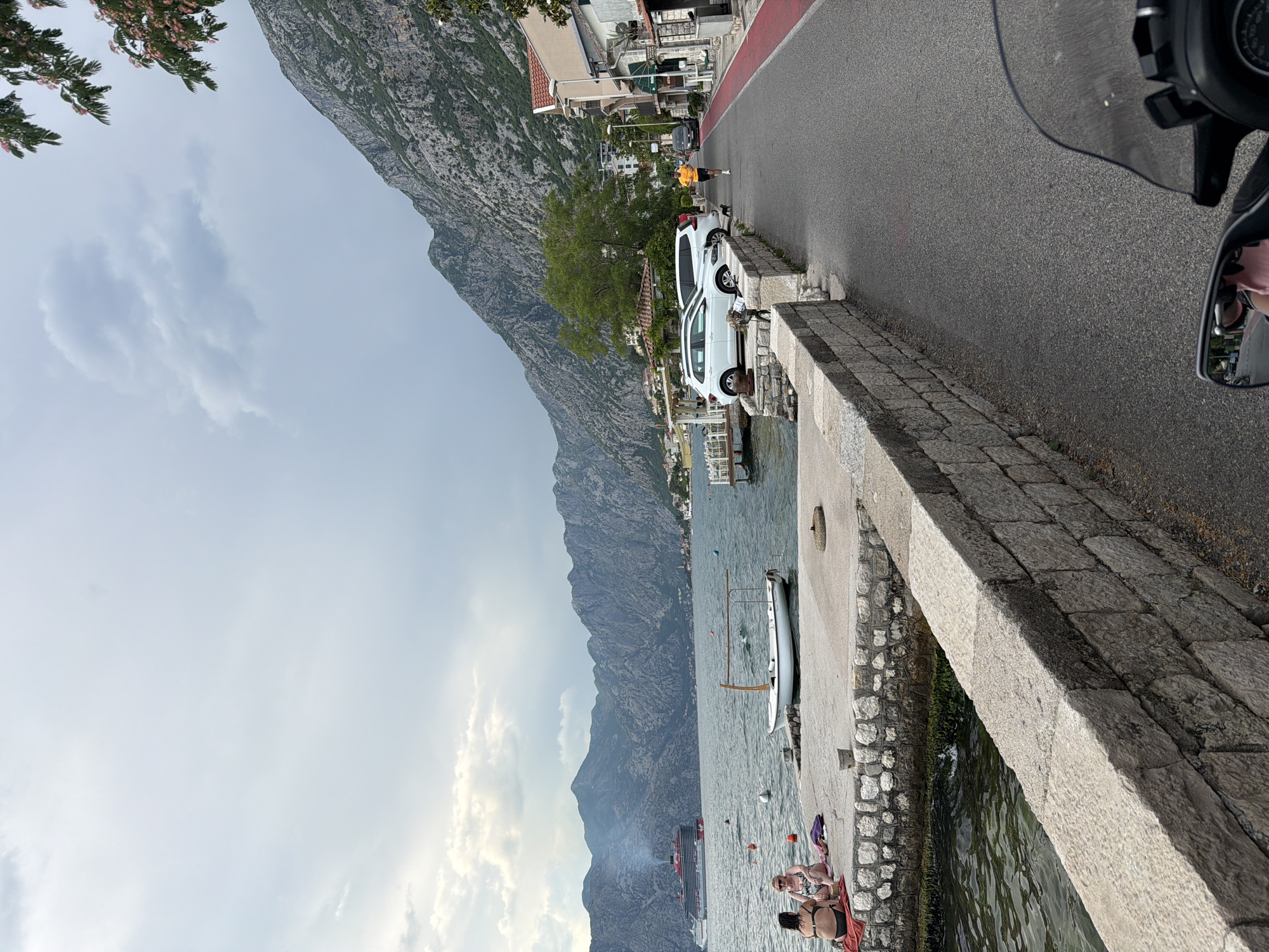 A high-angle, eye-level, medium shot captures a view along a paved road with a red lane marker, adjacent to a stone wall overlooking a body of water. In the distance, rugged mountains rise dramatically, their slopes dotted with houses and vegetation. A large cruise ship is visible on the water, and a small white boat is moored near a concrete pier.
In the foreground, two women are seated on a red towel on the concrete pier, facing the water. One woman is wearing a black bikini, and the other is in a patterned swimsuit. A small white boat with a wooden frame is tied to the pier.
On the road, a man in an orange t-shirt and black shorts is jogging. A black cat is walking near the side of the road, and two white cars are parked on the other side. Buildings line the right side of the road, with balconies and windows visible. The sky is overcast with patches of blue. The overall impression is a scenic coastal town with recreational activities taking place.
The scene is located in Dobrota, Montenegro, a town situated along the Bay of Kotor. The distinctive mountainous landscape and the presence of the bay are characteristic of this region.