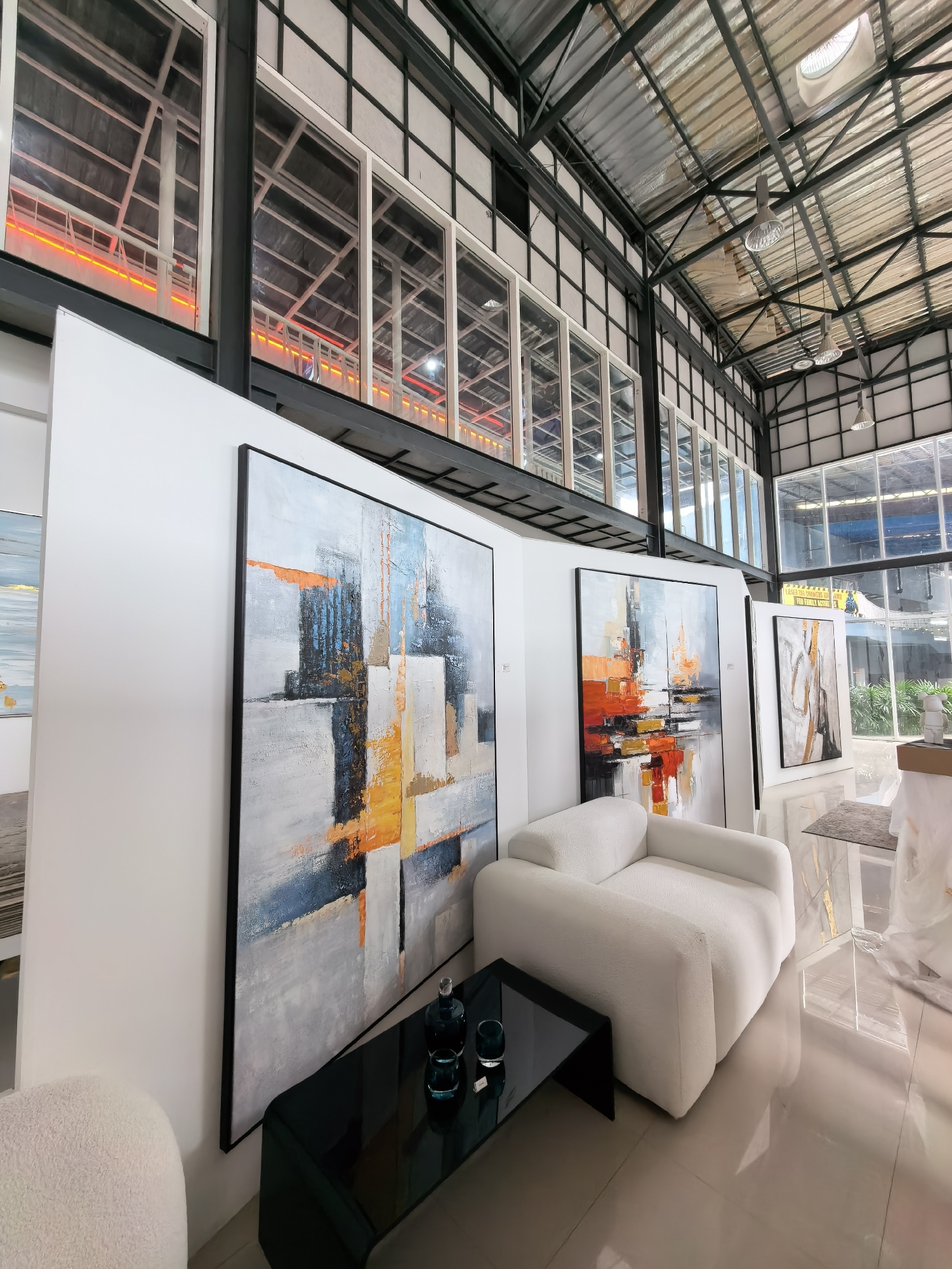 The image shows an interior view of an art gallery or showroom. The space has a high ceiling with exposed metal beams and corrugated metal roofing, suggesting an industrial or warehouse-like structure. Large windows run along the upper portion of the walls, allowing natural light to enter. Several large abstract paintings are displayed on white walls. In the foreground, a plush white armchair sits next to a sleek black coffee table, on which two dark glass vases are placed. Another abstract painting is visible on the wall behind the armchair, and further back, more artwork and furniture can be seen. The floor is tiled and highly reflective. The overall impression is a modern, minimalist space showcasing contemporary art.