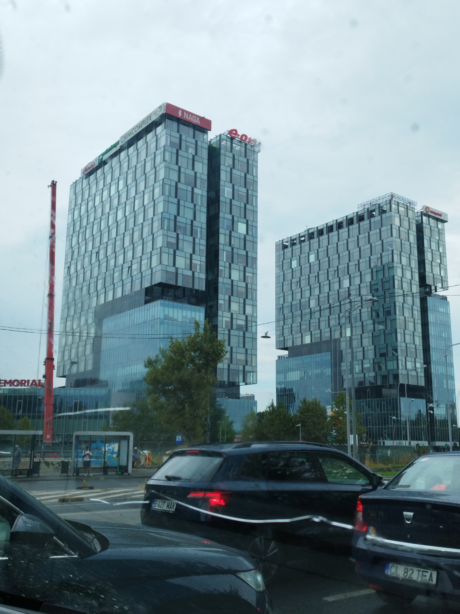 The image depicts a street scene in Bucharest, Romania, likely during daytime, under an overcast sky. The foreground features the hoods and windshields of two dark-colored cars, suggesting the photograph was taken from inside a vehicle. In the mid-ground, traffic is visible on a multi-lane road. A dark blue sedan, with license plate B 427 MAK, is stopped or moving slowly in the right lane. Another dark-colored vehicle is visible to the right, with license plate CL 82 TFA. Beyond the road, several modern, glass-facade buildings dominate the skyline. The tallest building, on the left, features prominent signage for "NAGA" and "CHIMCOMPLEX". To its right, another similar building displays "e.on" branding. Further to the right, a third building is partially visible with "computered" signage. The buildings are characterized by their grid-like arrangement of windows. Trees are interspersed between the road and the buildings, and a red construction crane is visible to the left of the "NAGA" building. A bus stop shelter is present on the sidewalk with a few individuals standing near it. The overall impression is that of a busy urban environment with significant modern architectural development.