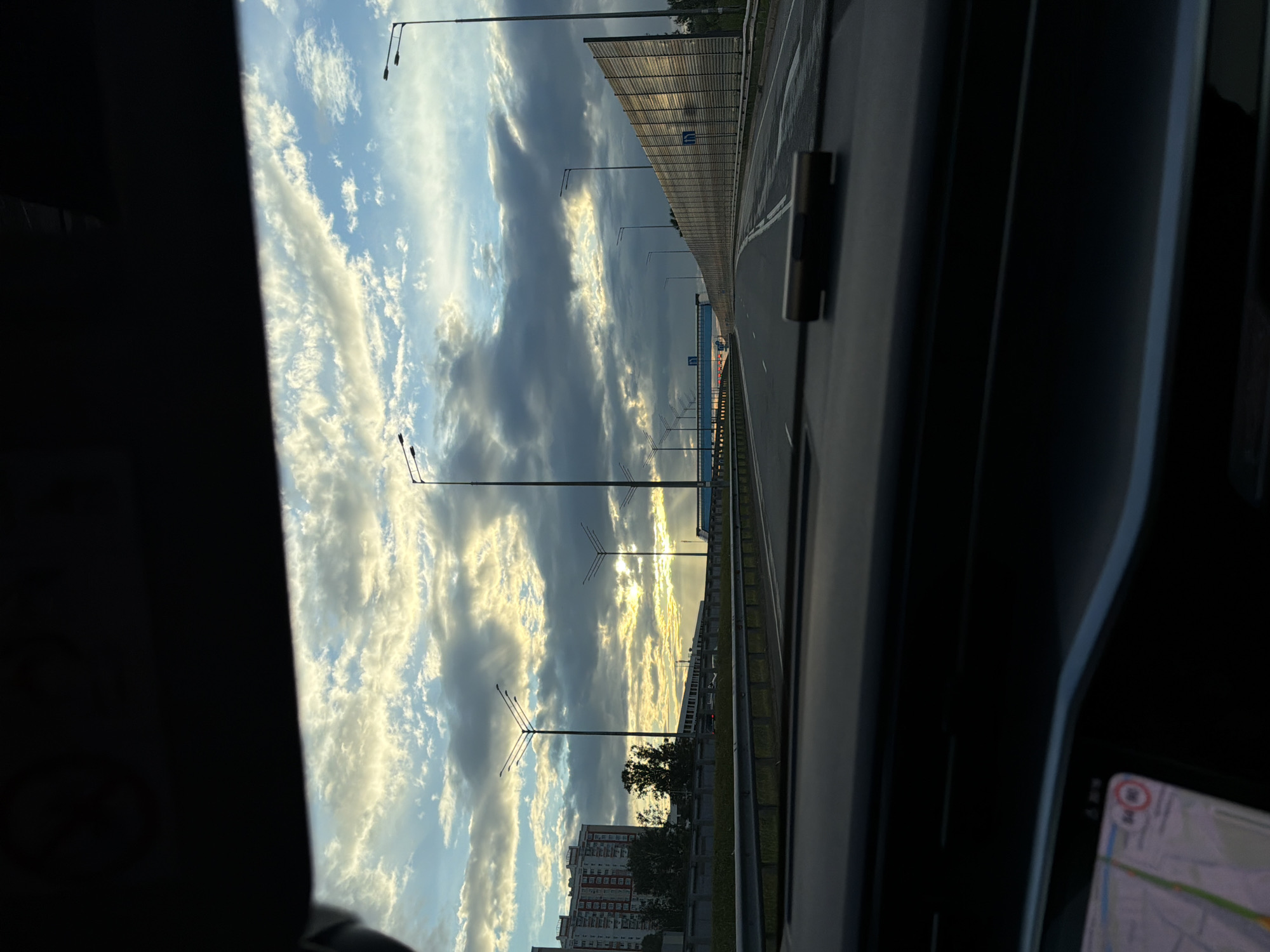 The image, taken from inside a vehicle, depicts a view of a highway in Khimki, Russia, during what appears to be sunrise or sunset. The sky is filled with dramatic clouds, with rays of sunlight breaking through in the distance. There are multiple streetlights lining the highway, and a sound barrier wall is visible on the right side of the road. In the background, a tall residential building is situated to the left, and an overpass with blue accents spans across the highway in the center. The foreground shows the dashboard of the car, including a navigational device displaying a map. There are no people or animals visible in the image.