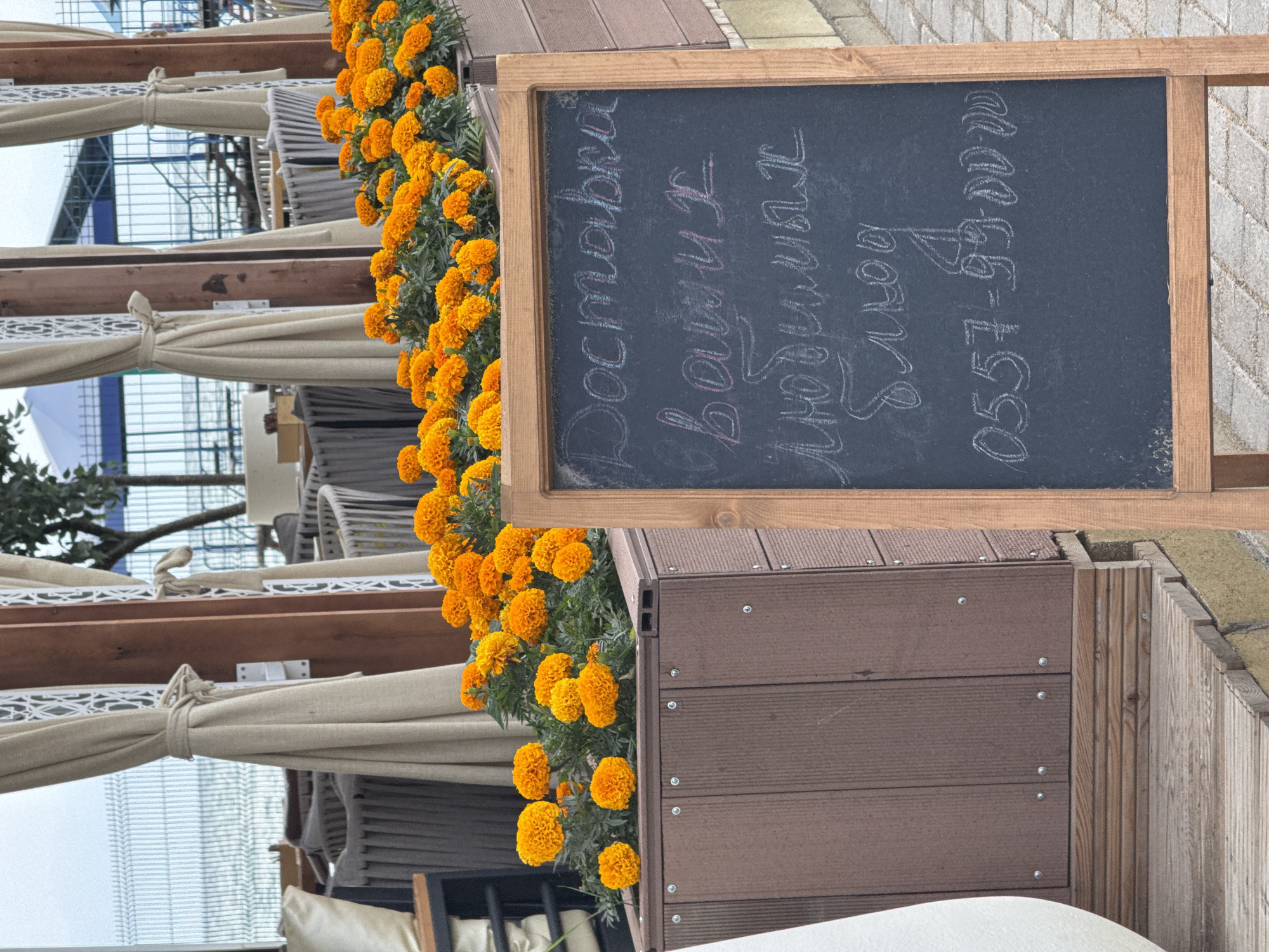 The image shows an outdoor seating area with orange flowers in a planter box. A chalkboard sign stands in front of the planter, displaying handwritten text in white chalk. The text appears to be in Russian and reads "Доставка ваших любимых блюд 0557-99-00-00". The scene is likely located in Sary-Oy, Kyrgyzstan, given the style of outdoor dining and the presence of a fence and water in the background, suggesting a proximity to a lake or sea. There are no people visible in the image.