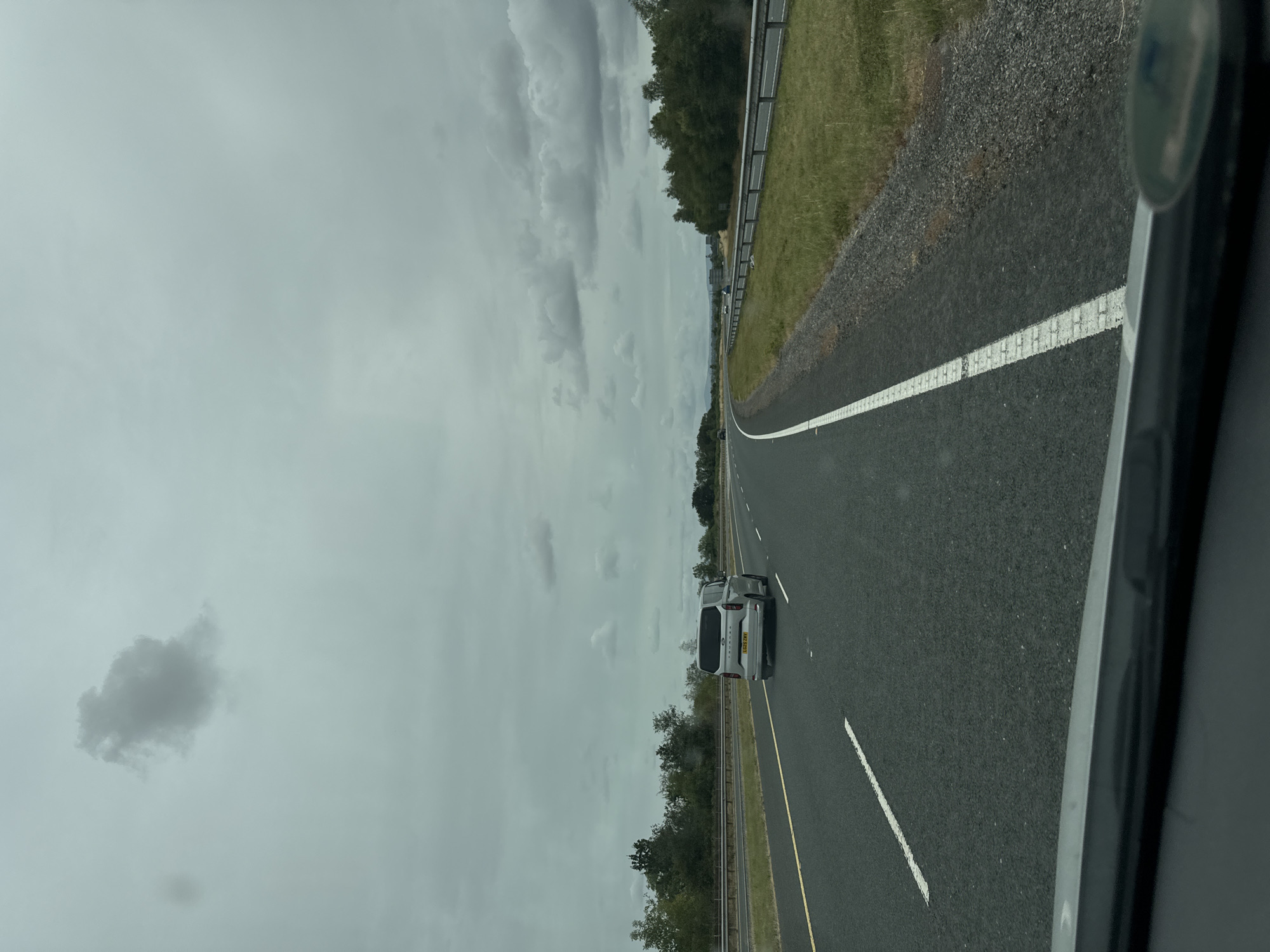 The image captures a view from a vehicle traveling on a highway, likely in or near Tinryland, Ireland, given the context. The sky is overcast with dark clouds, suggesting a gloomy day. The highway is paved asphalt with clear white lane markings and a yellow line marking the right edge of the lane. On the left side of the road, there is a metal guardrail separating the highway from a grassy verge and a line of trees. On the right side, the road curves slightly, with a grassy shoulder and a metal guardrail. A silver van is visible in the left lane ahead, with its license plate clearly visible. The foreground shows the edge of the vehicle's dashboard and windshield. The overall impression is one of a typical drive on a highway under cloudy skies in a rural setting.