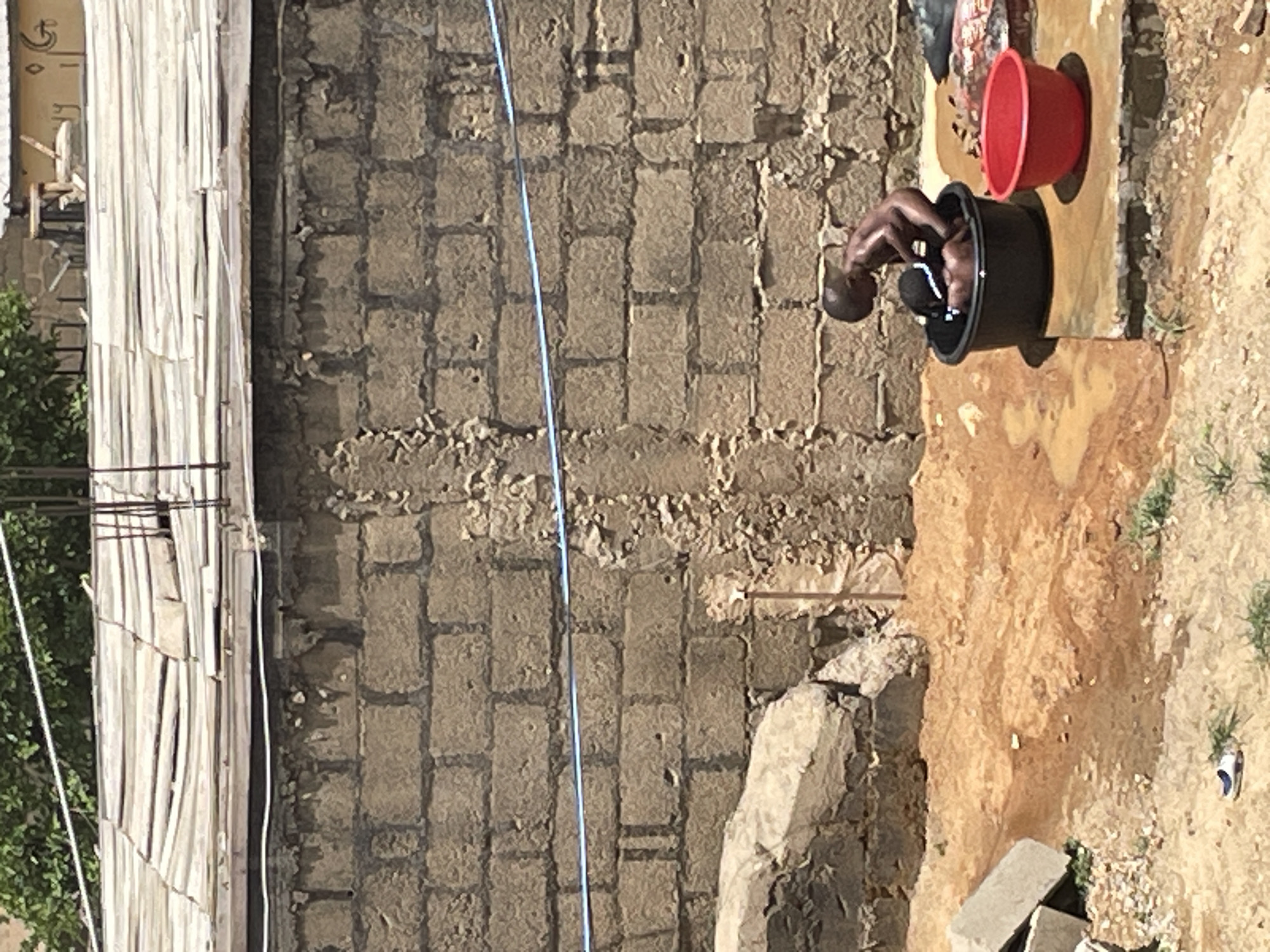 A man bathes outdoors in Jos, Nigeria. The man is bent over a black bucket, using a white cloth. A red bucket sits nearby on a concrete surface stained with spilled water. A cinder block wall forms the backdrop, with a blue cord running vertically along it. To the left, weathered planks are visible. The ground is dry and sandy, with scattered vegetation.
