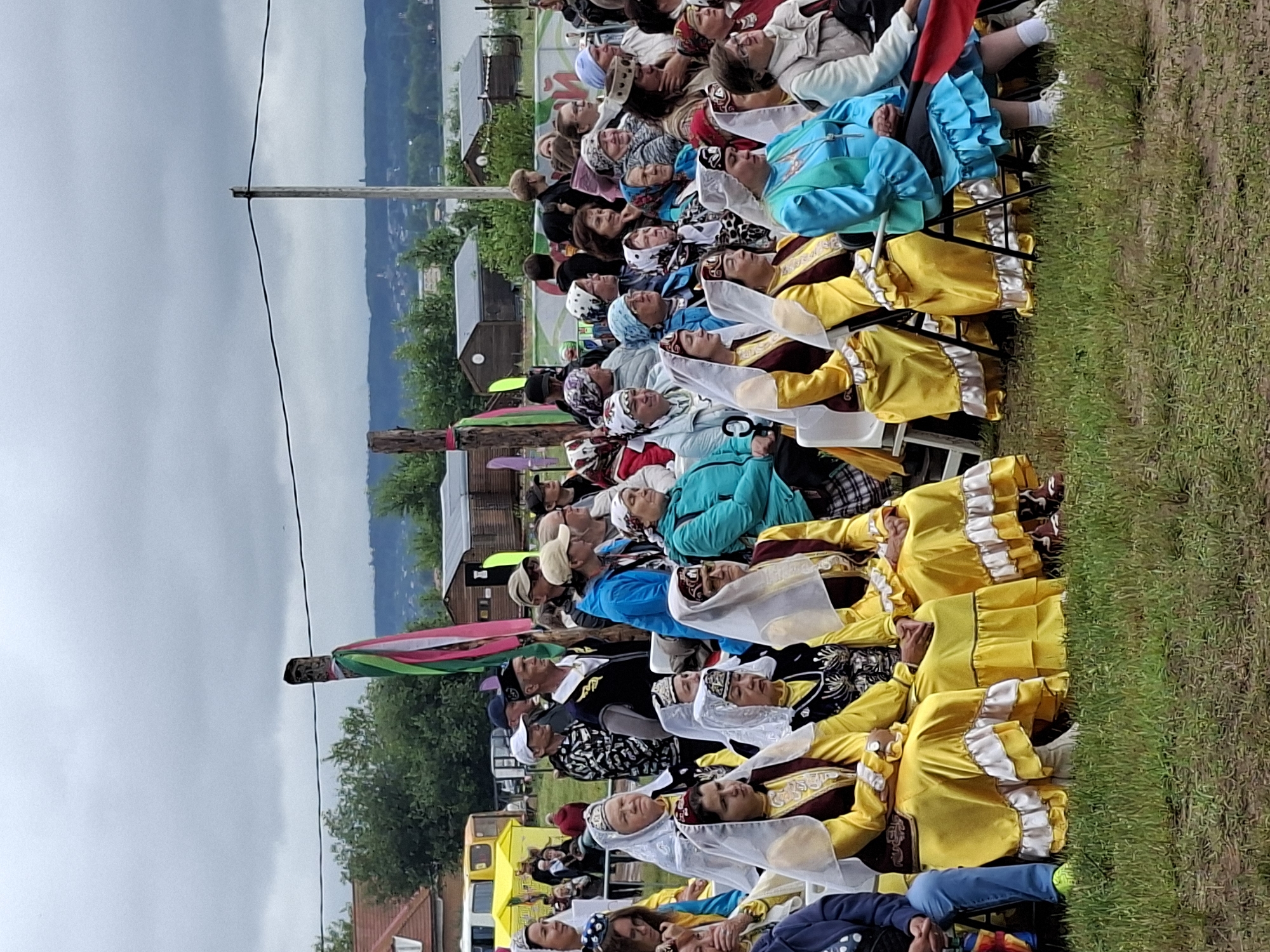 This image captures an outdoor gathering in Votkinsk, Russia, likely a cultural event. A crowd of people, predominantly women, are seated on chairs outdoors, facing towards the right side of the frame. Many of the women are wearing traditional, brightly colored clothing, primarily in yellow and burgundy, often adorned with intricate embroidery and headscarves. Some are wearing white veils over their heads.
In the background, several wooden structures, resembling small sheds or houses, are visible. There are also trees and a body of water, possibly a river or lake, in the distance. A yellow bus is parked on the left side of the image, and a power line stretches across the upper portion of the scene. Two wooden poles with colorful ribbons are planted in the ground, suggesting a ceremonial or festive purpose for the gathering. The sky is overcast with heavy clouds. The overall atmosphere appears to be one of a community event or festival, with an emphasis on traditional attire and cultural celebration.
