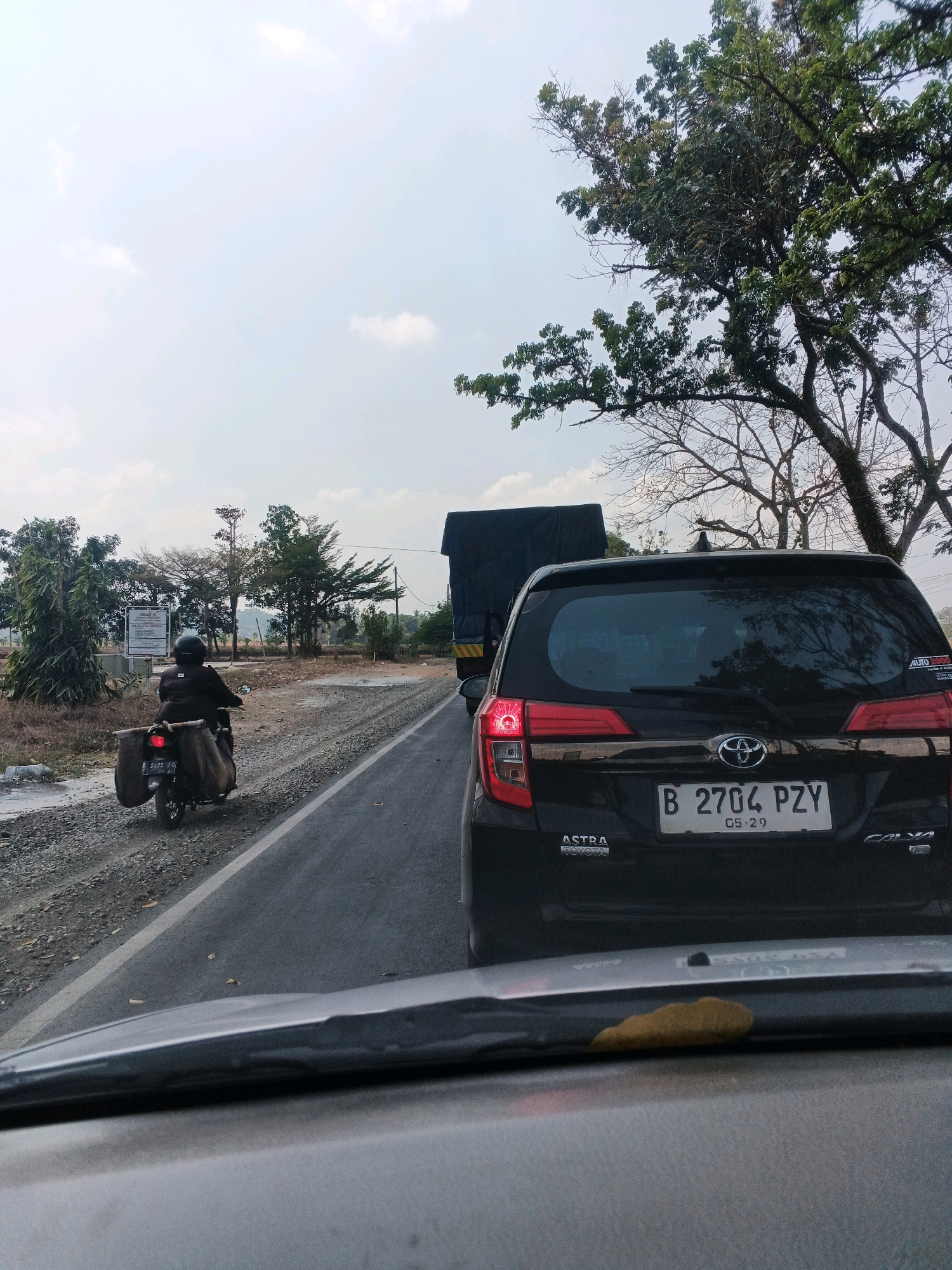 A dark-colored car is stopped on a paved road in Kedungpring, Indonesia, seen from behind and slightly above. The car has a license plate that reads "B 2704 PZY 05-29". A truck is visible in front of the car, also stopped. To the left of the road, a person on a motorcycle with large bags attached to the back is also stopped. The road appears to be a two-lane street with a gravel shoulder on the left. Trees and vegetation are visible on both sides of the road, and the sky is overcast. A sign is visible in the background to the left, with some text in Indonesian. The scene suggests a traffic delay or congestion on a rural or semi-rural road.