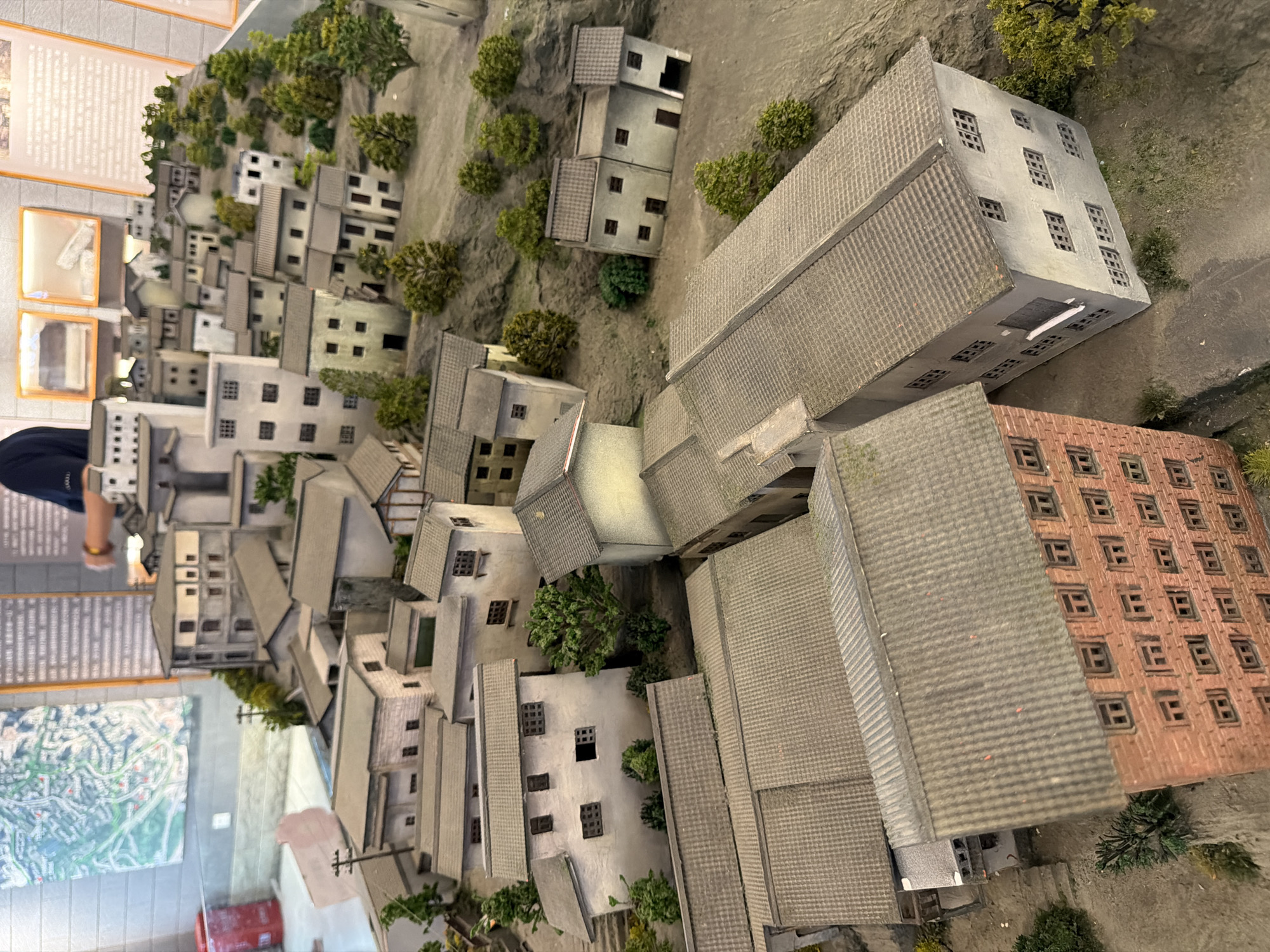 The image displays a detailed diorama of a densely packed urban landscape, likely representing a historical or present-day scene in Yuzhong District, China. The diorama features numerous multi-story buildings with tiled roofs, varying in color from light gray to beige and some with visible brickwork. The architecture suggests a traditional Chinese urban style, with narrow streets and clustered houses. Miniature trees and vegetation are strategically placed to simulate a hilly terrain. In the background, educational displays are visible, including a map and informational texts mounted on the wall. A partially visible person, wearing a dark shirt, is standing behind the diorama, interacting with the display. The diorama appears to be well-lit, highlighting the intricate details of the miniature city.