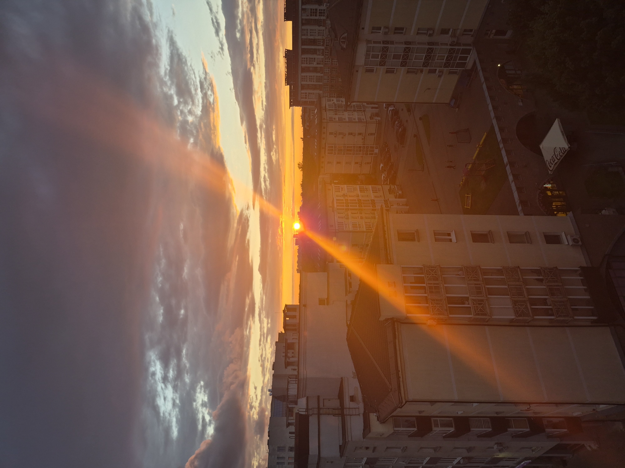 The image depicts a vibrant sunset over a cityscape, likely in Minsk, Belarus, given the architectural style of the buildings which are typical of post-Soviet urban development. The sun, a bright orange orb, is low on the horizon, casting strong, radiant beams of light that cut through the dusky sky and illuminate the apartment buildings. The sky is a dramatic blend of dark, brooding clouds and warm, fiery hues of orange, pink, and yellow, creating a stark contrast. Several multi-story residential buildings dominate the foreground and midground. Their facades are a mix of beige and off-white, with numerous windows and balconies. Some buildings have visible architectural details such as external ladders, air conditioning units, and decorative railings on balconies. In the foreground, a prominent building features a distinctive red-tiled roof. Further back, more apartment blocks stretch into the distance. A children's playground with swings and a small soccer goal is visible in a courtyard area between buildings, as is a Coca-Cola branded awning extending from a ground-floor establishment. A few cars are parked in designated areas. The overall impression is one of urban life transitioning into evening, bathed in the dramatic glow of a setting sun.