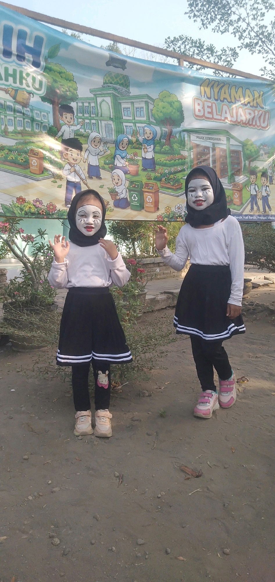 In the city of Plemahan, Indonesia, two young girls stand before a large banner depicting a school setting with cartoon children and buildings. Both girls are wearing black hijabs and white long-sleeved shirts. Their faces are painted with white makeup and black markings around their eyes and mouths, resembling clown or theatrical makeup. The girl on the left wears a black skirt over black leggings with a cartoon bunny appliqué on the left leg. The girl on the right wears a black pleated skirt over black leggings. Both are wearing light-colored sneakers. The banner features text that appears to read "NYAMAN BELAJARKU" and "IH AHAKU," suggesting an educational theme. The environment appears to be outdoors, with dirt ground, some potted plants, and trees visible in the background, under a bright sky.