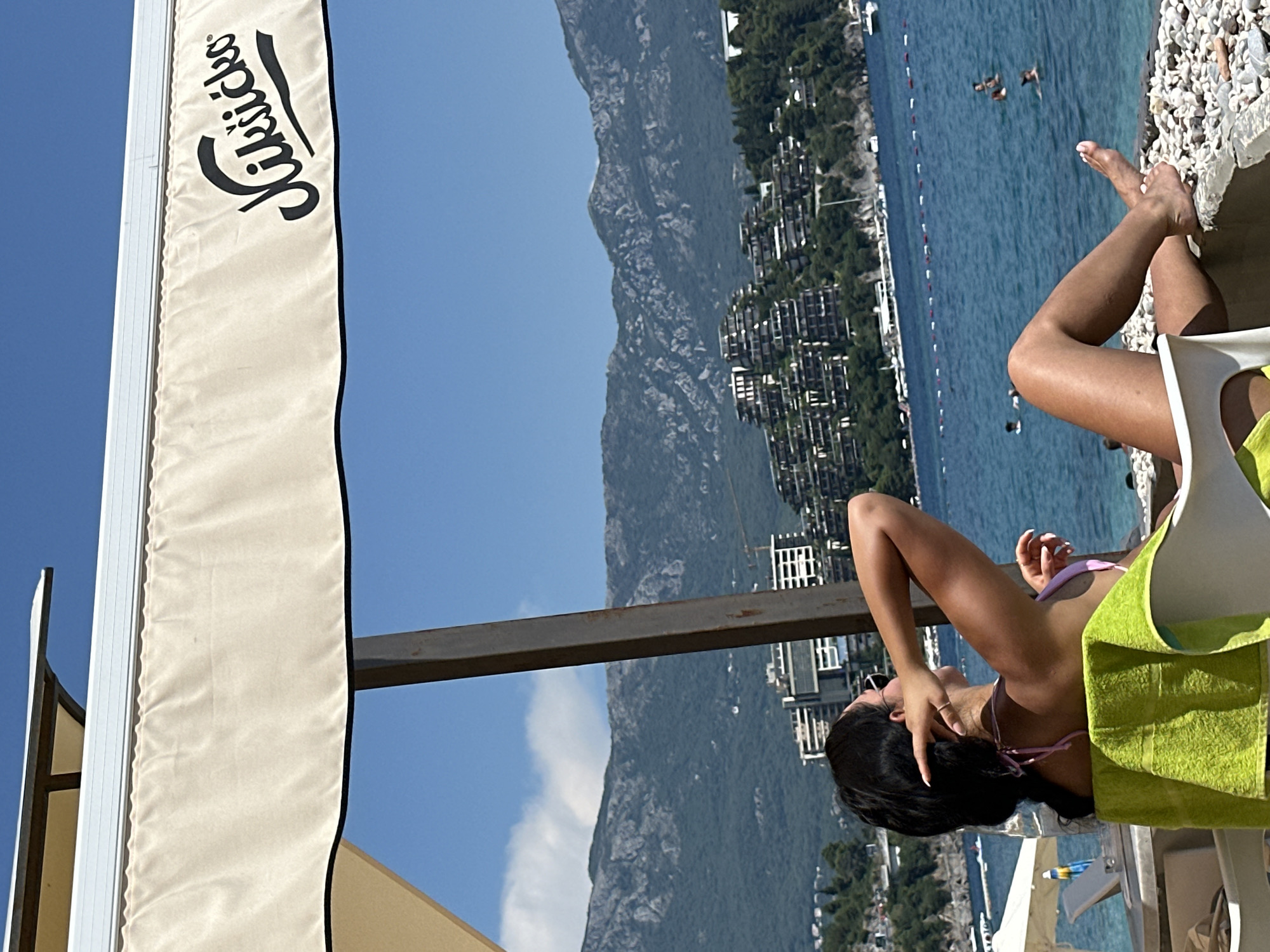 This image captures a serene scene at a beach resort. A woman, dressed in a pink bikini, is relaxing on a lounge chair. She has dark hair and appears to be enjoying the sun, with one hand resting on her head. A bright green towel is draped over the chair beside her.
The background reveals a picturesque coastal view. A tranquil blue sea stretches out, dotted with a few boats and a buoys marking swimming areas. Along the shoreline, a densely built-up area with white buildings and lush green trees climbs up a steep, rocky mountain. The sky is clear and blue, suggesting a sunny day.
To the left, a section of a cream-colored awning is visible, with a black trim. A black logo, appearing to be a stylized text in cursive, is prominently displayed on the awning. The overall atmosphere is one of relaxation and vacation, typical of a summer day at a beach destination. The time of day appears to be daytime, likely late morning or afternoon, given the bright sunlight. The location is contextually identified as Budva, Montenegro.