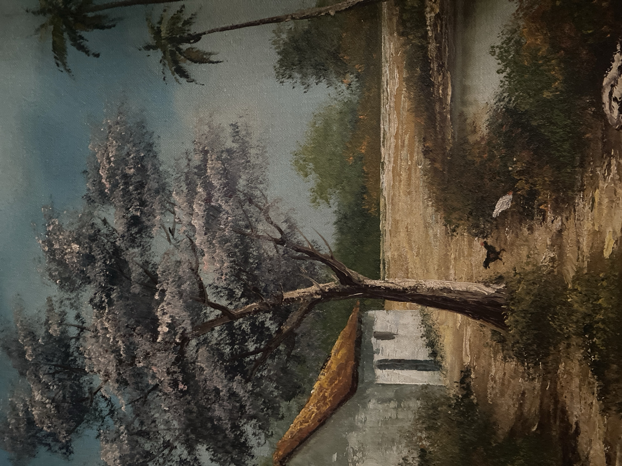 The image is an oil painting of a landscape. In the upper left corner are two palm trees against a blue-grey sky. A large tree with light purple-grey foliage dominates the left side of the painting.
To the right of the tree, a tan vertical structure that may be a wall or a bluff is visible. At the base of the structure is a light grey building with a dark brown roof. A dark brown tree branch extends from the foliage on the left over the building and towards the right.
The right side of the painting depicts a wooded area with a path or waterway running vertically along the edge of the wall. There are two birds, one white and one black, in the wooded area, and another dark bird in flight. The exact location within the city of Tinaquillo, Venezuela is not discernable from the content of the painting.