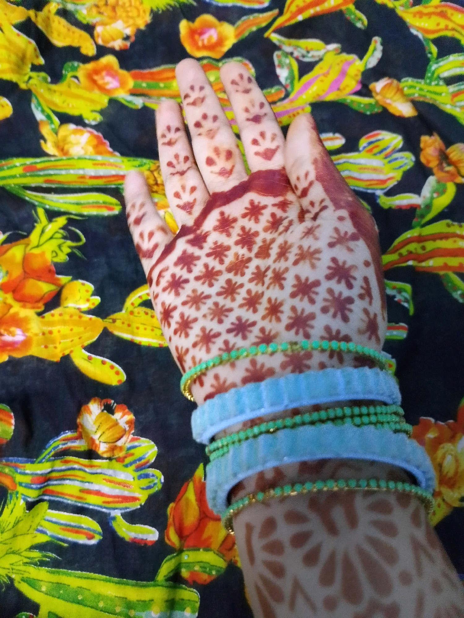 A person's left hand, adorned with henna designs and bangles, is held up against a vibrant, dark-colored fabric featuring a colorful floral and abstract pattern. The henna designs on the fingers appear as small, simplified faces with dots for eyes and curved lines for mouths. The palm of the hand displays a more intricate pattern of small, star-like or floral shapes in a reddish-brown hue. Stacked on the wrist are three bangles: the topmost is a wide, light blue fabric bangle, followed by two thinner bangles comprised of small, iridescent green beads with gold trim. The background fabric is rich with imagery, including yellow and orange floral elements and abstract shapes in yellow, green, red, and blue, evoking a sense of lively, perhaps festive, decoration. This image was taken in Thane, India.