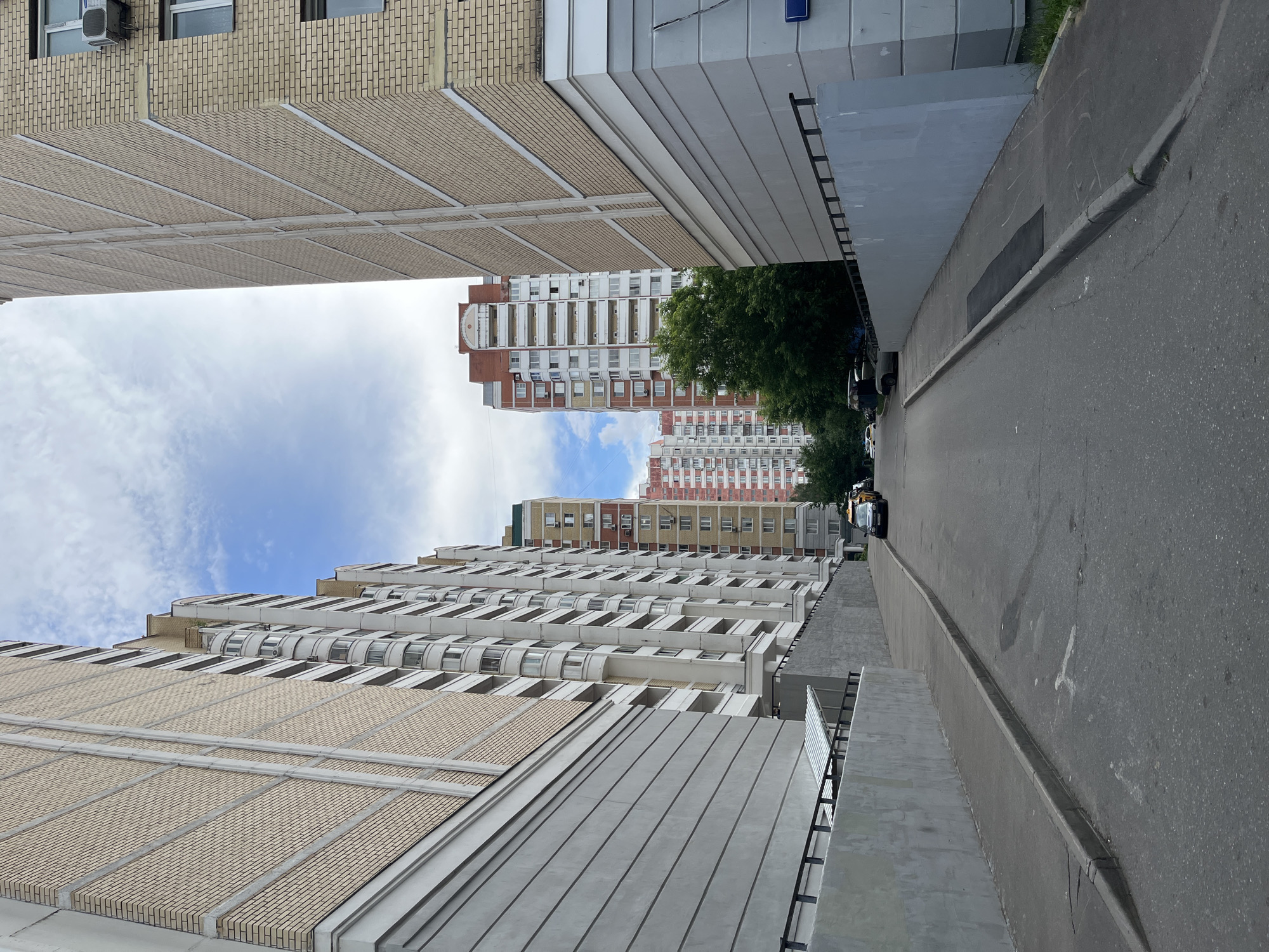 The image depicts a street scene in Moscow, Russia, with tall, modern apartment buildings flanking a paved road. The buildings on either side are constructed with a light-colored brick exterior and feature numerous windows and balconies. On the left, the building has a prominent grey concrete base and a sloping walkway with a metal railing. On the right, another apartment building has a similar light brick facade and a grey concrete lower section. In the background, more residential buildings of varying heights and architectural styles are visible, some with red and white exteriors. Green trees line parts of the street, and the sky above is partly cloudy. Several cars are parked or driving on the road, including a yellow taxi. The overall impression is that of a typical urban residential area.