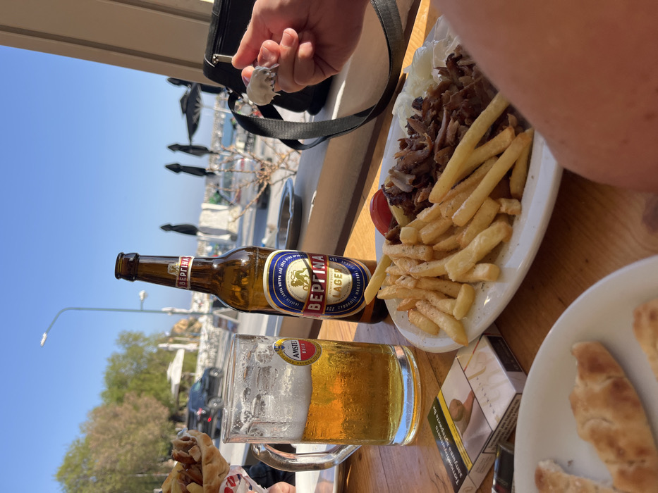 The image shows a table in an outdoor restaurant in Limenaria, Greece, with coordinates 40.626, 24.576. On the table there is a glass of Amstel beer, a bottle of "BEΠΙΝA Lager" beer, a plate of french fries and sliced meat with a tomato on top, a package of cigarettes and some pita bread. A person's right arm and hand can be seen holding a white object. The environment is sunny and bright, and there is a window behind the scene showing a view of the sea. The table is wooden and the seats are wooden chairs. There are two black umbrellas in the background. The person in the image is not visible in full. The image is taken at a slightly elevated angle.