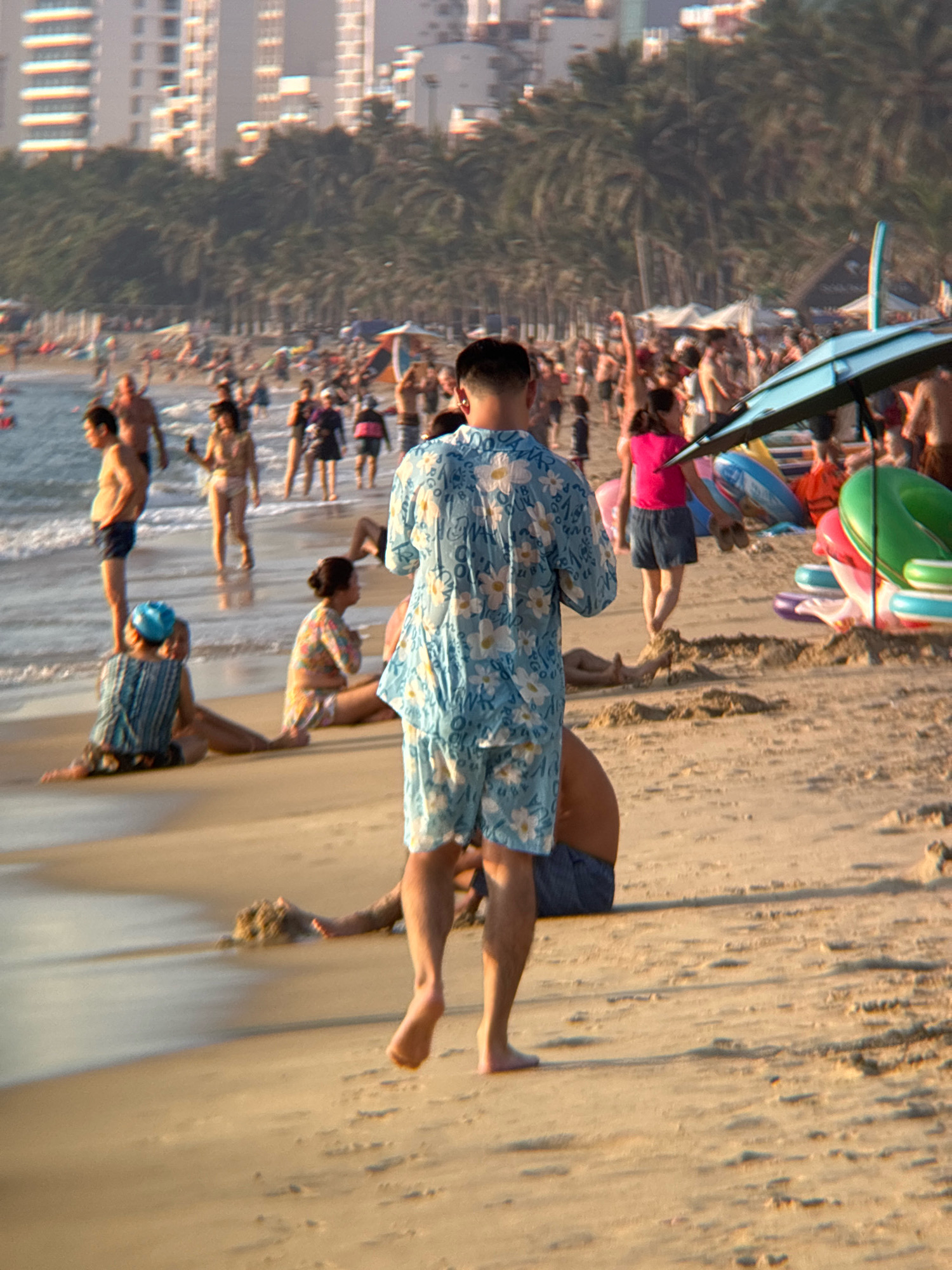 The image depicts a crowded beach scene in Nha Trang, Vietnam, likely during the day, given the bright sunlight. In the foreground, a person wearing a light blue shirt with a floral pattern and blue shorts is walking away from the camera, their back turned. To their left, a person in a blue and white striped shirt and dark shorts, wearing a blue swim cap, is sitting on the sand with their legs extended towards the water. To the right of this seated person, another individual in a floral-patterned top and shorts is also sitting on the sand. The background is filled with numerous people engaged in various beach activities, some walking along the shore, others gathered in groups. The ocean is visible with gentle waves lapping the shore. In the distance, a line of palm trees separates the beach from a backdrop of modern buildings. Beach umbrellas and inflatable toys are scattered across the sand, suggesting a recreational atmosphere.