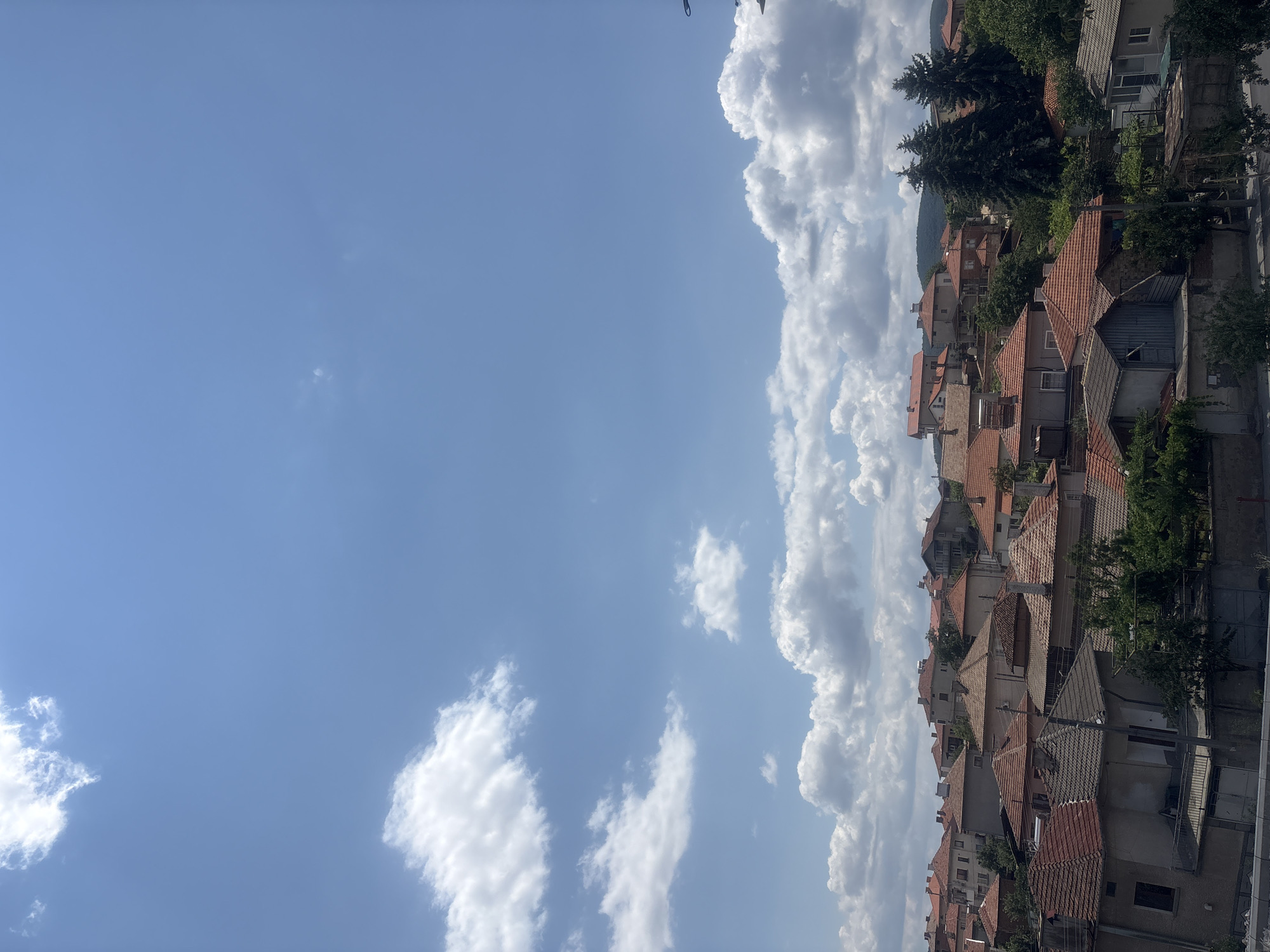 The image depicts a view of a residential area in the city of Strelcha, Bulgaria. The scene is dominated by a clear blue sky with scattered white clouds. Below the sky, a dense cluster of houses with red-tiled roofs fills the frame. The architecture appears to be traditional, with varying sizes and designs of buildings. Some houses have visible windows, balconies, and chimneys. Lush green trees are interspersed among the houses, adding a touch of nature to the urban landscape. The overall impression is a peaceful, sun-drenched day in a Bulgarian town.