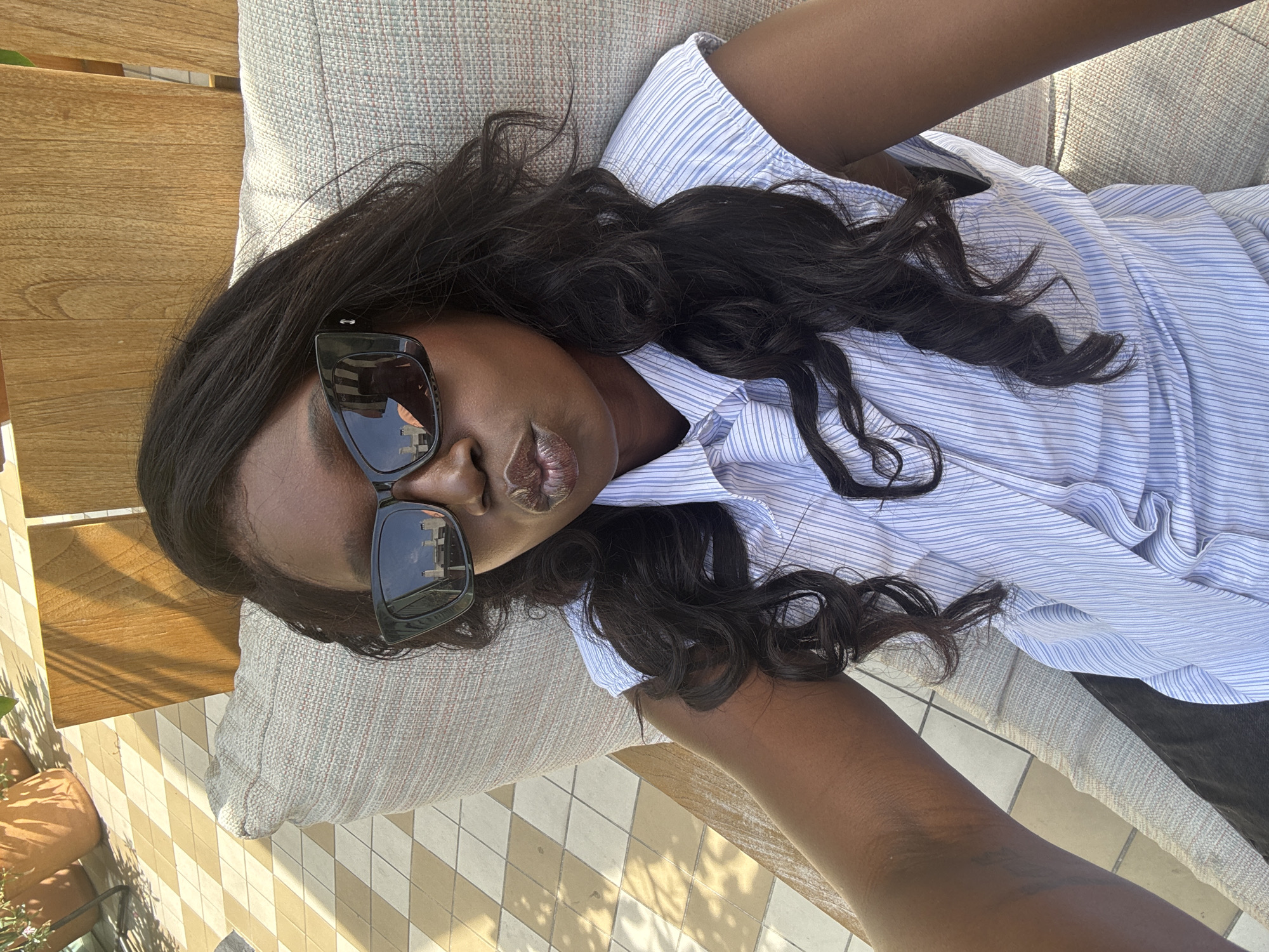 The image, taken in West Hollywood, United States of America, features a person relaxing outdoors in bright daylight. The subject, appearing to be a woman with a dark complexion, is lying on what looks like an outdoor lounge chair or bench, covered with a light-colored, textured cushion. She is looking directly towards the camera, possibly taking a selfie, with her lips subtly puckered.
She wears a light blue and white striped button-down shirt and large, dark sunglasses. The sunglasses have reflective lenses, showing a blurred reflection of a building or urban environment. Her long, dark, wavy hair is spread around her head and shoulders, resting on the cushion and her shirt. Her left arm is visible, extending towards the lower right of the frame.
The background includes a distinctive tiled floor with an alternating light and dark beige diamond or checkerboard pattern. Strong shadows are cast across the floor, indicating a sunny day. A wooden structure, possibly part of the furniture or a nearby railing, is visible behind the cushion. In the far left, glimpses of terracotta-colored pots or planters can be seen. The overall scene suggests a leisurely moment on a patio, balcony, or poolside area. There is no visible text in the image.
