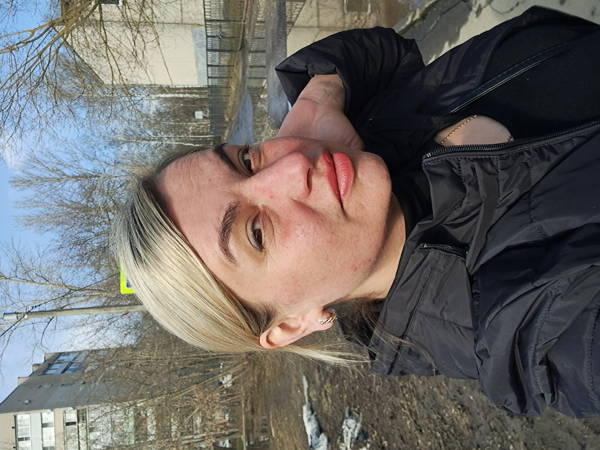 A woman with blonde hair is standing outdoors, likely in Elektrostal, Russia. She is wearing a black puffer jacket and a gold chain necklace is visible. The background includes bare trees, a building, a fence, and patches of snow on the ground.