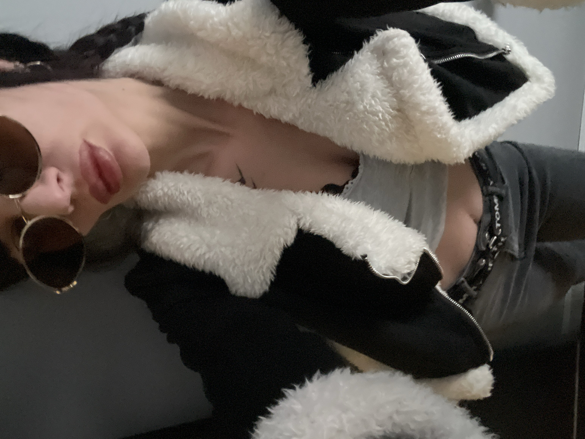A woman with sunglasses is photographed indoors. She is wearing a black and white jacket. She is also wearing a grey tank top and grey pants. A metal chain is visible around her waist. The location of the photo is not discernible based on the image.