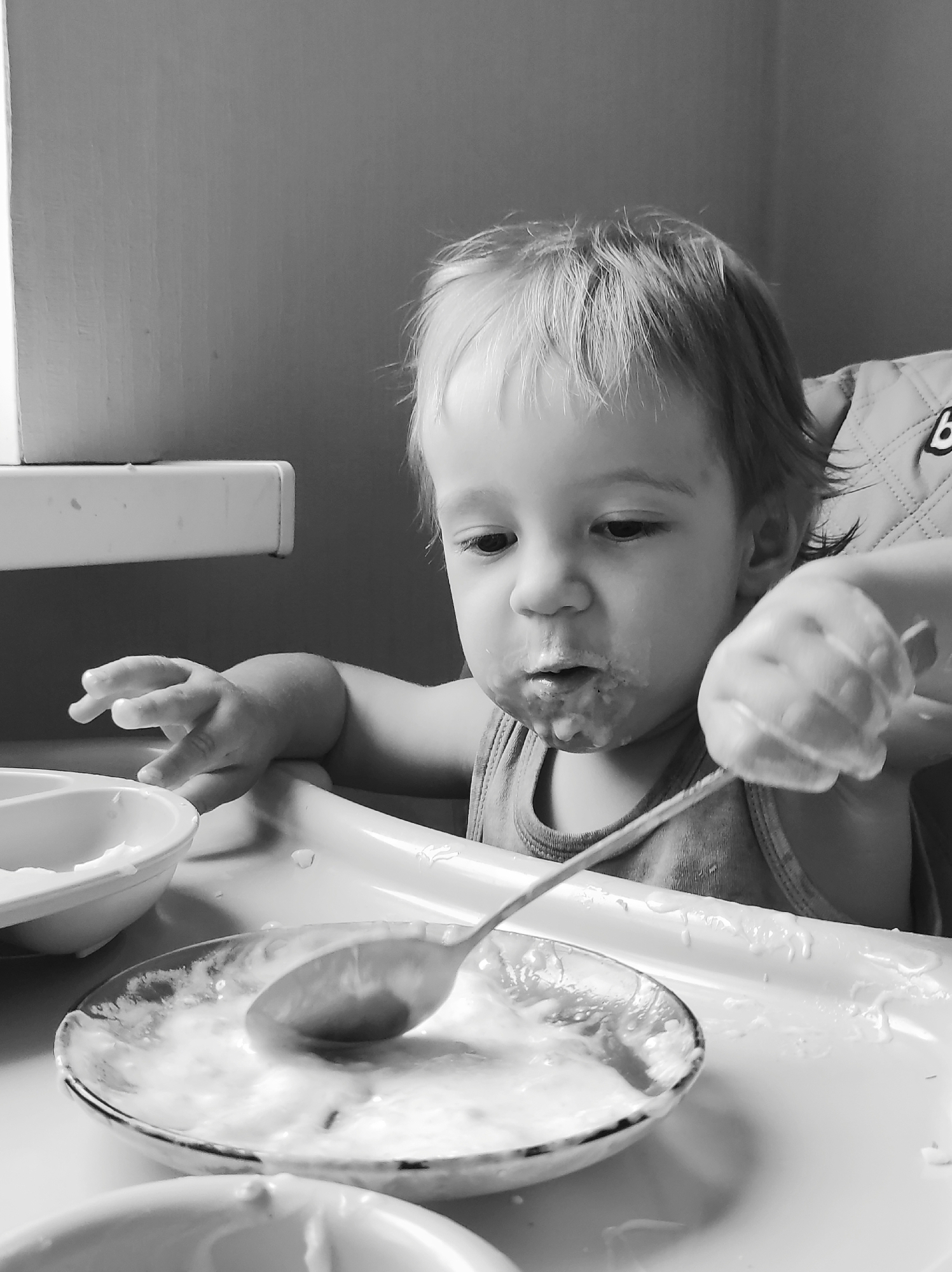 A black and white, medium shot photograph shows a young child, likely a toddler, sitting in a high chair. The child has light-colored hair and is looking down at a plate in front of them. Their face and chin are smeared with food. In their right hand, the child is holding a spoon, which is partially submerged in the food on the plate. The plate is round and appears to be made of glass, with a patterned rim. Beside it, to the left, is a white bowl, also containing food. The high chair tray is white and shows traces of food splatters. The background consists of a wall and a ledge or shelf. The overall scene suggests a mealtime, likely breakfast or lunch, given the lighting which appears to be natural daylight. The child's expression is focused on the food, with a hint of messiness and perhaps a touch of determination to feed themselves. The setting is domestic, likely a home kitchen or dining area. There is no visible text in the image.