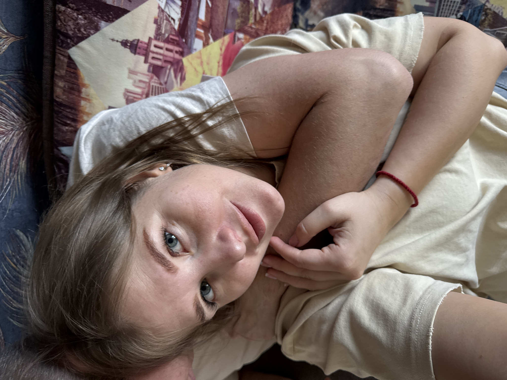 A young woman lies on a patterned cushion in what appears to be an indoor setting. She is wearing a light-colored t-shirt and a red bracelet. Her hair is brown, and her eyes are blue. She looks directly at the camera, with her hand gently touching her face. The background includes a cushion with a design that resembles a cityscape or architectural structure. There is not enough information to provide a specific location in Chaikovsky, Russia.
