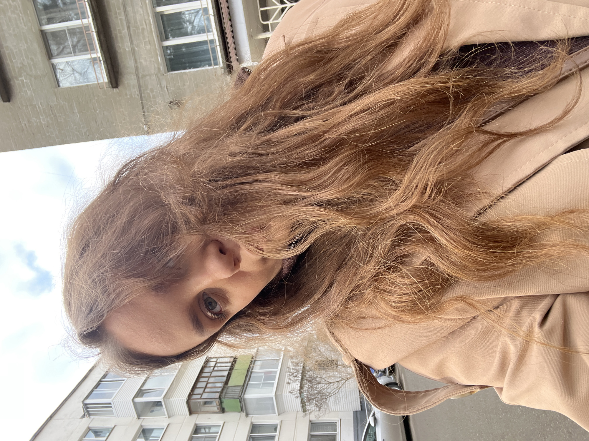 A young woman is the subject of a low-angle selfie. She is fair-skinned with light brown hair. She wears a tan coat. The background features buildings with windows and balconies. The location is Chaikovsky, Russia.