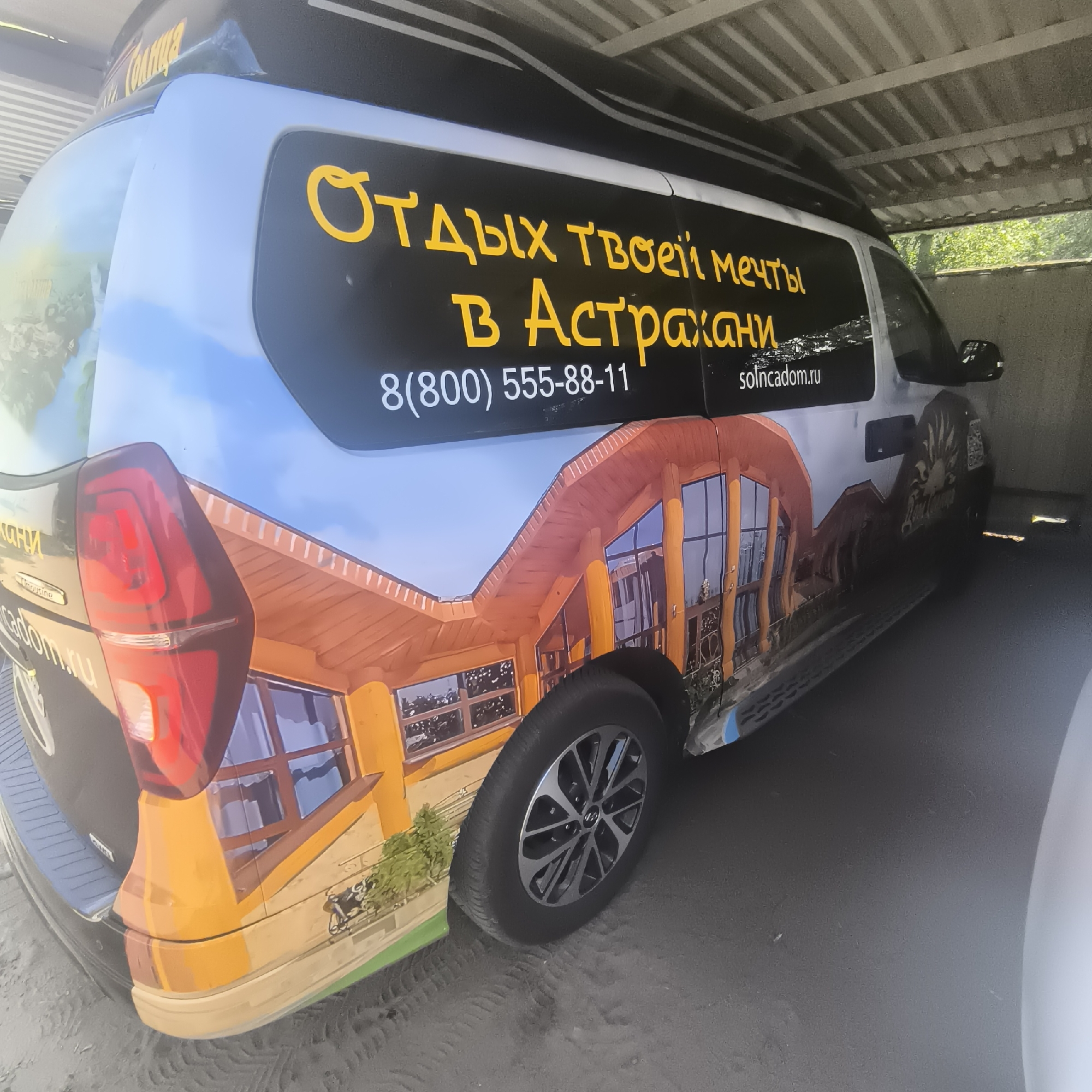The image shows the side of a vehicle parked under a covered structure. The vehicle is a light blue van with a large graphic on its side. The graphic depicts a stylized house with a bright orange roof and walls, featuring several windows. Above the house graphic, there is a black banner with yellow text that reads "ОТДЫХ ТВОЕЙ МЕЧТЫ В АСТРАХАНИ", which translates to "Vacation of Your Dreams in Astrakhan". Below this text, a phone number "8(800) 555-88-11" and a website "solincadom.ru" are visible. The vehicle also has a red taillight on the left side and a side mirror on the right. The ground appears to be sandy or dusty, with tire tracks visible. The structure above the vehicle is a corrugated metal roof supported by beams. The overall scene suggests the vehicle is in an outdoor parking area, possibly a campsite or resort. The image does not contain any people, nor does it provide any specific location information within the city of Malyy Baklaniy, Russia.