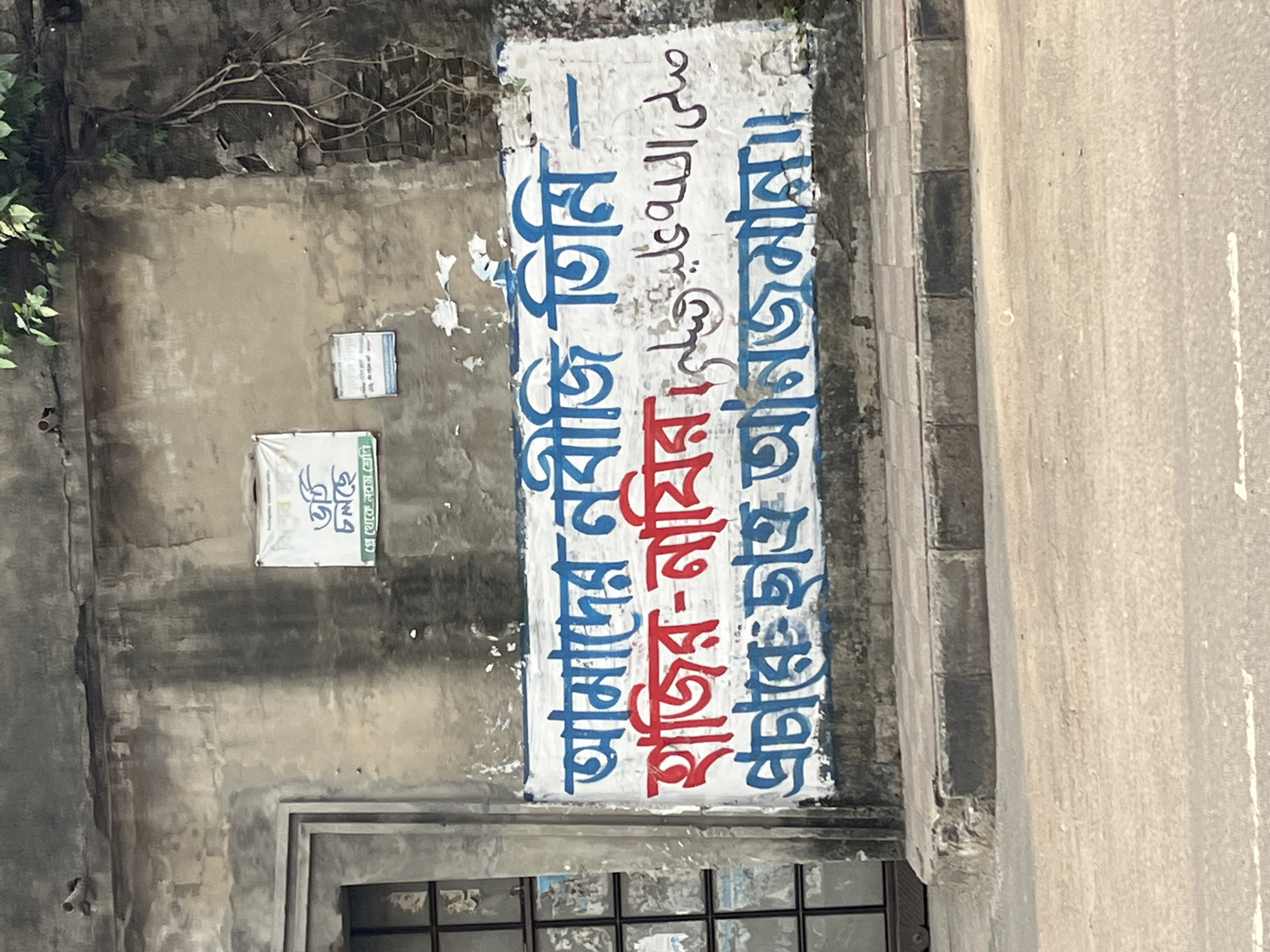 The image shows a weathered wall in Dhaka, Bangladesh. On the wall are two signs, one partially obscured. The larger sign is a white rectangular panel painted with blue and red Bengali script, and Arabic script in black. The script translates to: "Our prophet is present", "Hajir Nazir", and "Student Anjuman". The text also includes the Arabic invocation "Sallallahu Alaihi Wasallam" (Peace and blessings of Allah be upon him). Above and to the left of the sign are other smaller signs. Below the signs, a glimpse of a window can be seen. To the right is a section of paved sidewalk and a road.