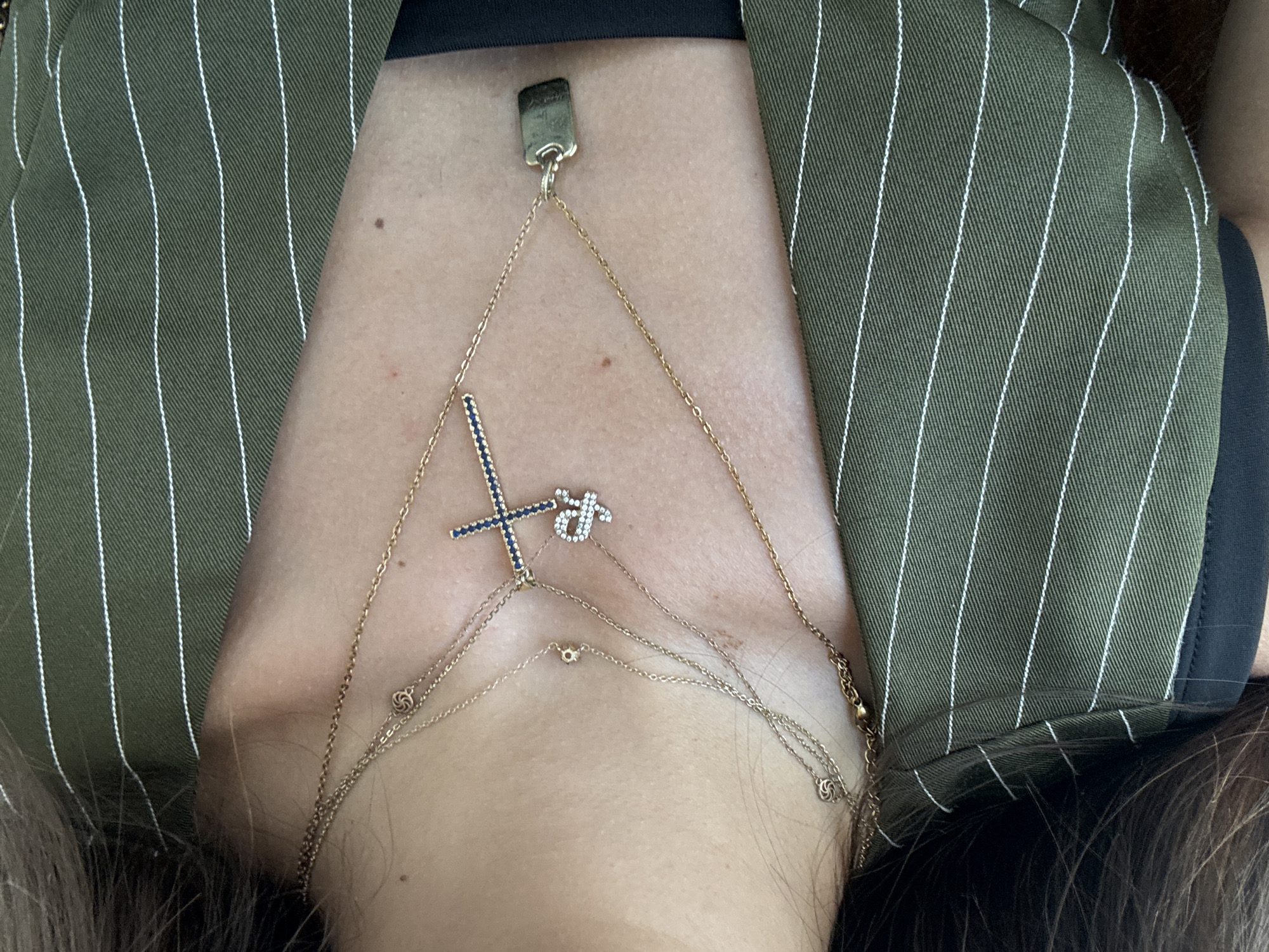 This media file shows a close-up of a person's chest wearing ornate gold necklaces. One necklace drapes down from the collarbone, featuring a pendant that appears to be a cross embellished with blue and clear stones, and a small, stylized letter "fr" also adorned with clear stones. The chains are layered and delicate, with small circular charms and intricate designs along their lengths. The person is wearing a dark top beneath a green and white pinstripe jacket. The setting appears to be indoors, with natural lighting. There is no discernible text in the image.