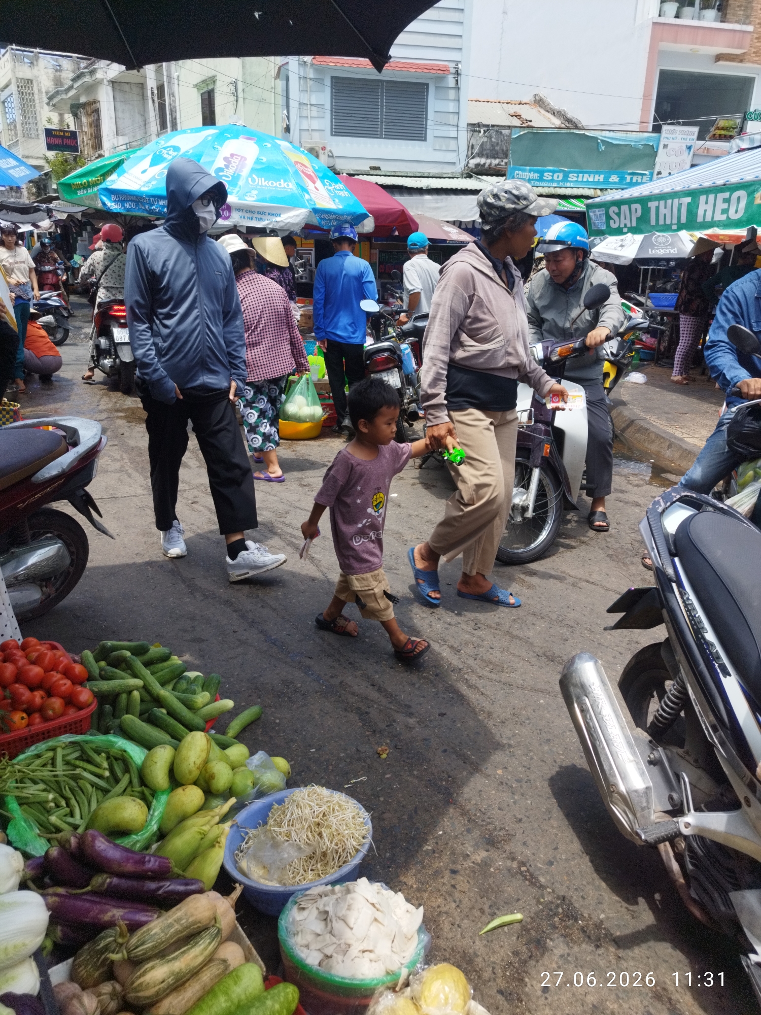 The image depicts a busy outdoor market scene in Phan Thiết, Vietnam. In the foreground, a variety of fresh produce is displayed, including tomatoes, cucumbers, okra, eggplants, corn, and bean sprouts. Several motorcycles are parked throughout the market.
People are seen walking and interacting. A person wearing a blue hoodie and a white mask is walking towards the viewer. A child in a purple shirt is walking away from the viewer, holding a small green toy. An adult in a light-colored hoodie and tan pants is walking in the same direction as the child, looking towards the child. Another adult in a blue shirt is seated on a motorcycle. Several other individuals are visible in the background, suggesting a crowded market atmosphere.
The environment appears to be a street market with temporary stalls and awnings providing shade. Buildings are visible in the background, some with signs. The ground is paved and appears wet in places, indicating it may have recently rained or is a common occurrence in the area. The overall impression is one of daily commerce and activity within a Vietnamese urban setting.