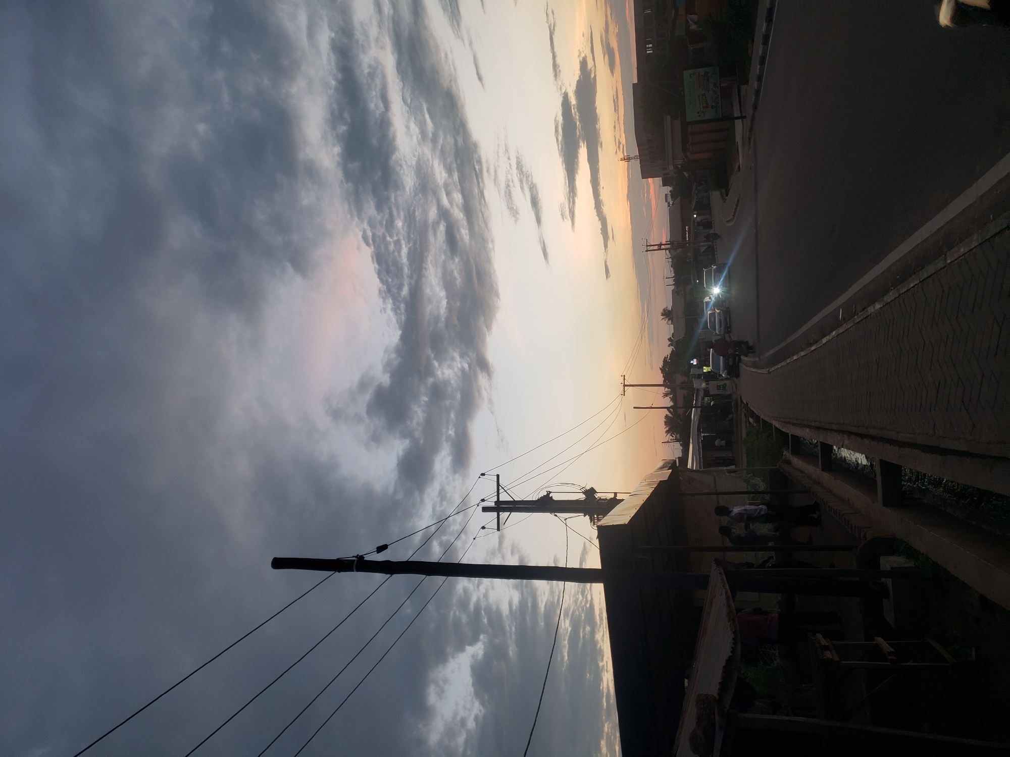This image captures a street scene at dusk in Narayi, Nigeria. The sky is filled with dramatic, dark grey clouds, with hints of pink and orange from the setting sun visible near the horizon. Power lines stretch across the frame, supported by utility poles, including one prominent wooden pole on the left and a metal one with crossbars further down the street.
On the left side of the road, there are makeshift structures with corrugated metal roofs and open sides, suggesting a market or roadside stalls. A few individuals are standing near these stalls, partially obscured by shadow. A concrete drainage channel runs alongside the sidewalk.
The asphalt road curves gently to the right, with several vehicles visible. Two cars with their headlights on are approaching, their beams cutting through the fading light. A motorcycle can also be seen. On the right side of the road, buildings of varying heights line the street, with some appearing to be commercial establishments. A billboard with colorful graphics is mounted on a wall. The overall atmosphere is subdued due to the low light conditions of dusk.