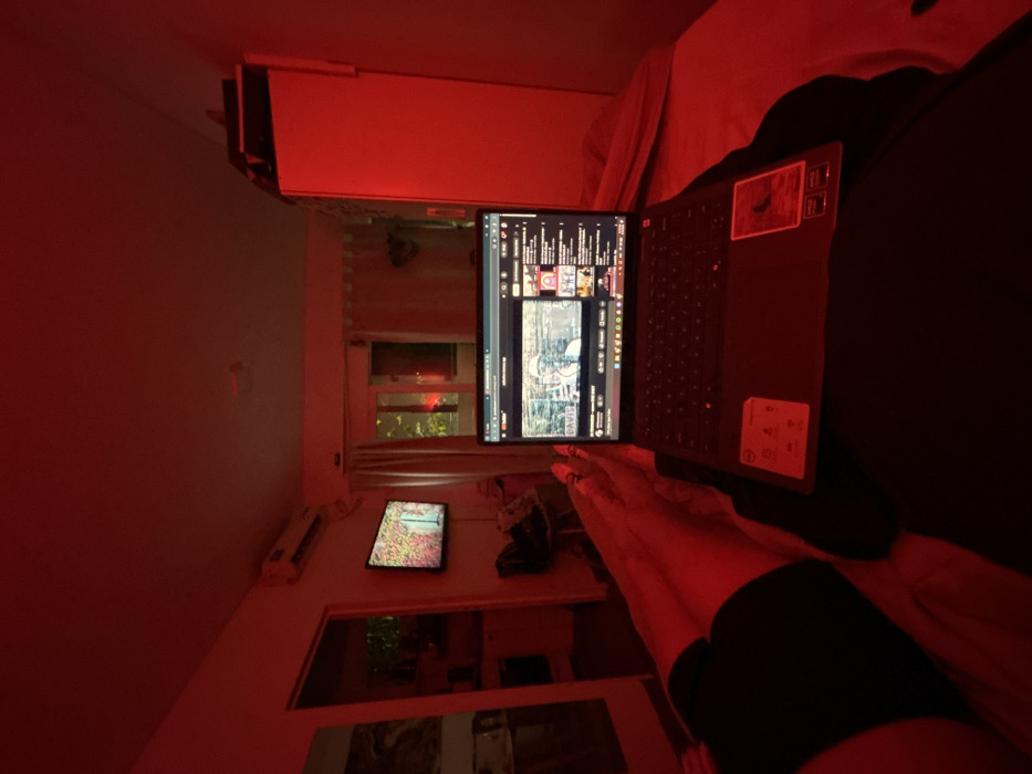 The scene depicts the interior of a residential apartment room in Buenos Aires, Argentina, illuminated almost entirely by a strong red light source. The time is nighttime, with a dark exterior visible through a window.
In the immediate foreground, the bare legs of an individual, wearing dark shorts, are extended, resting beneath an open dark-colored laptop computer. The laptop screen displays a YouTube interface, actively playing a video with the title "DAVIS" partially visible, alongside a sidebar of recommended videos. The laptop rests on a dark, unidentifiable surface, likely the person's lap or bedding.
In the midground, a flat-screen television is mounted on a light-colored wall, displaying a colorful image resembling foliage. Above the television, a white split-type air conditioning unit is affixed to the wall. To the right of the television, a window with closed light-colored curtains reveals the dimly lit outdoors, including tree branches and a distant red light source. To the far right, a tall, white wardrobe or cabinet is partially visible. To the left, an open doorway leads to an adjacent room, possibly a kitchen or hallway, where a large mirror or reflective surface is present on the left wall. The walls of the room are light-colored, but fully saturated by the red illumination. No specific district or address within Buenos Aires can be determined from the visual cues.